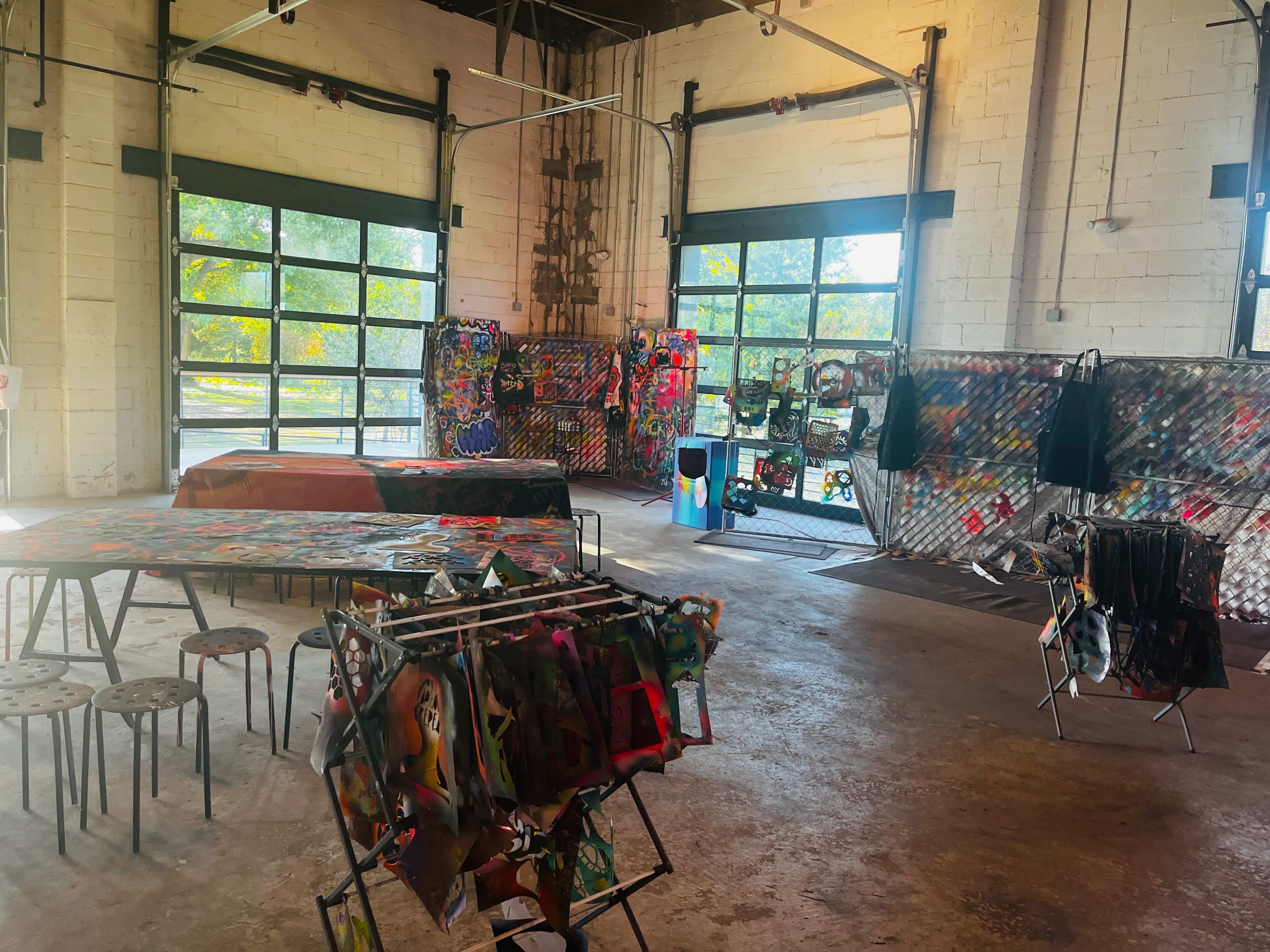 An industrial-style art space with tables displaying artwork and walls decorated with colorful pieces.