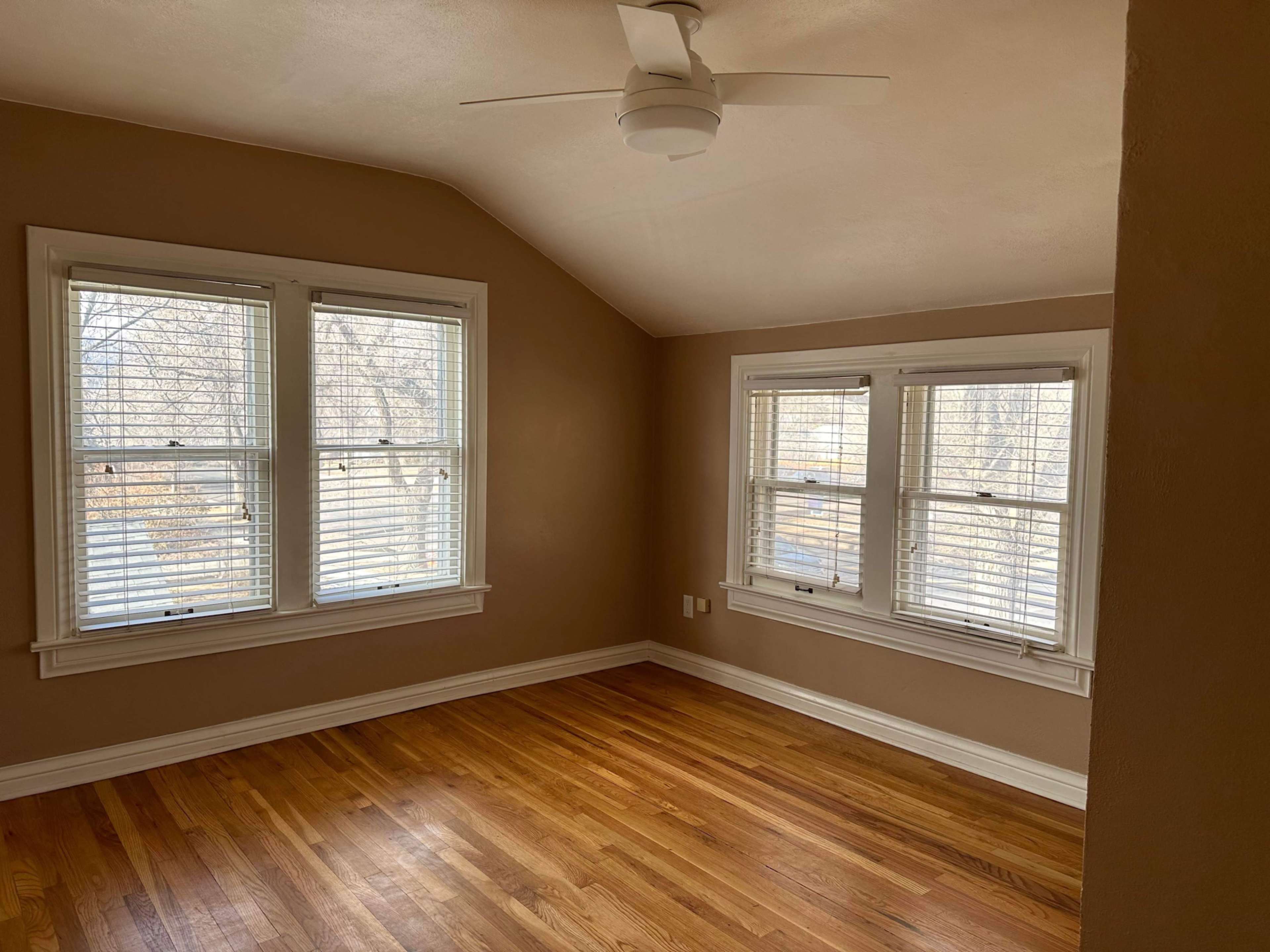 An empty room with sloped ceilings, featuring two sets of windows with blinds and hardwood flooring.