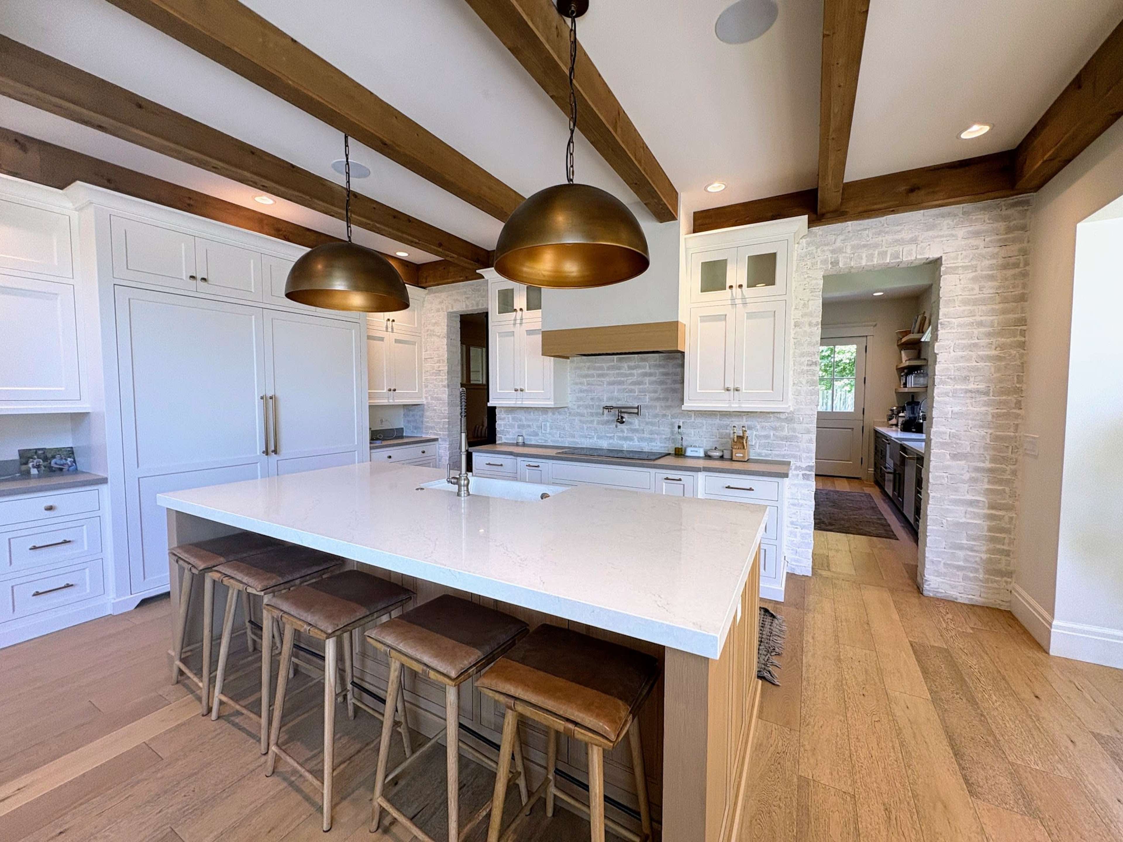 The kitchen features a large island with a white countertop, wooden beams on the ceiling, and brass pendant lights above.