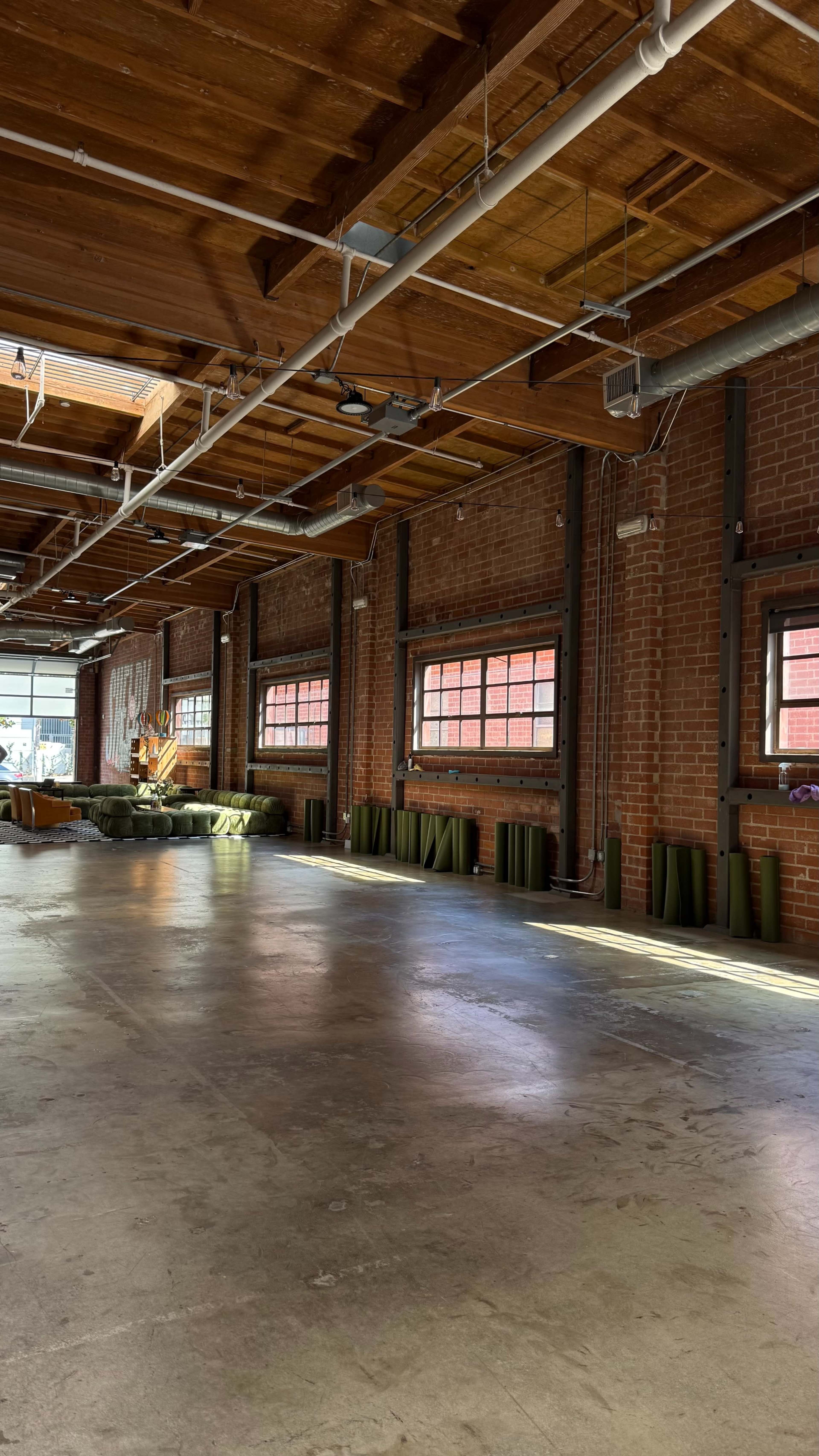 The image shows an empty industrial space with exposed brick walls, large windows allowing natural light, and concrete flooring.