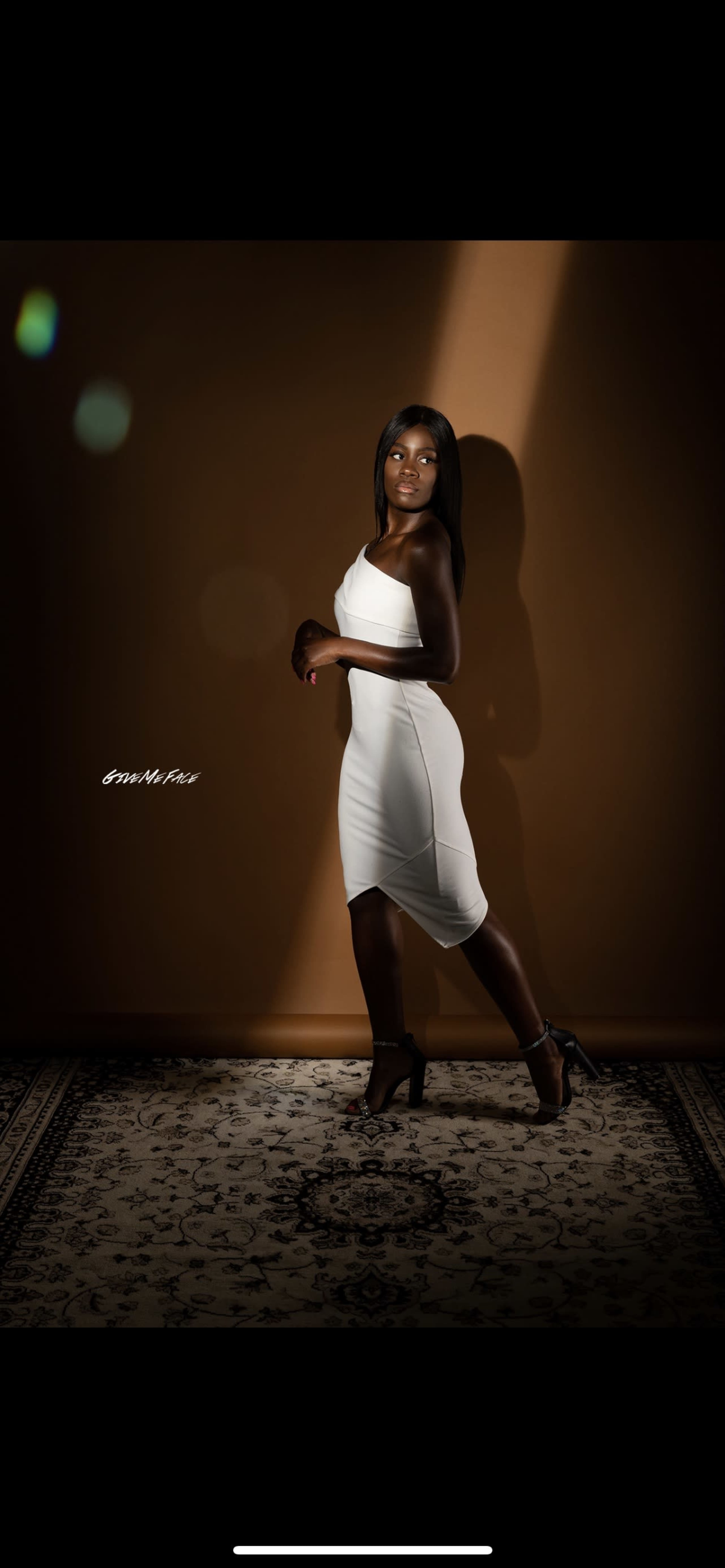 A person in a fitted white dress poses against a brown wall, with a spotlight illuminating part of the scene.