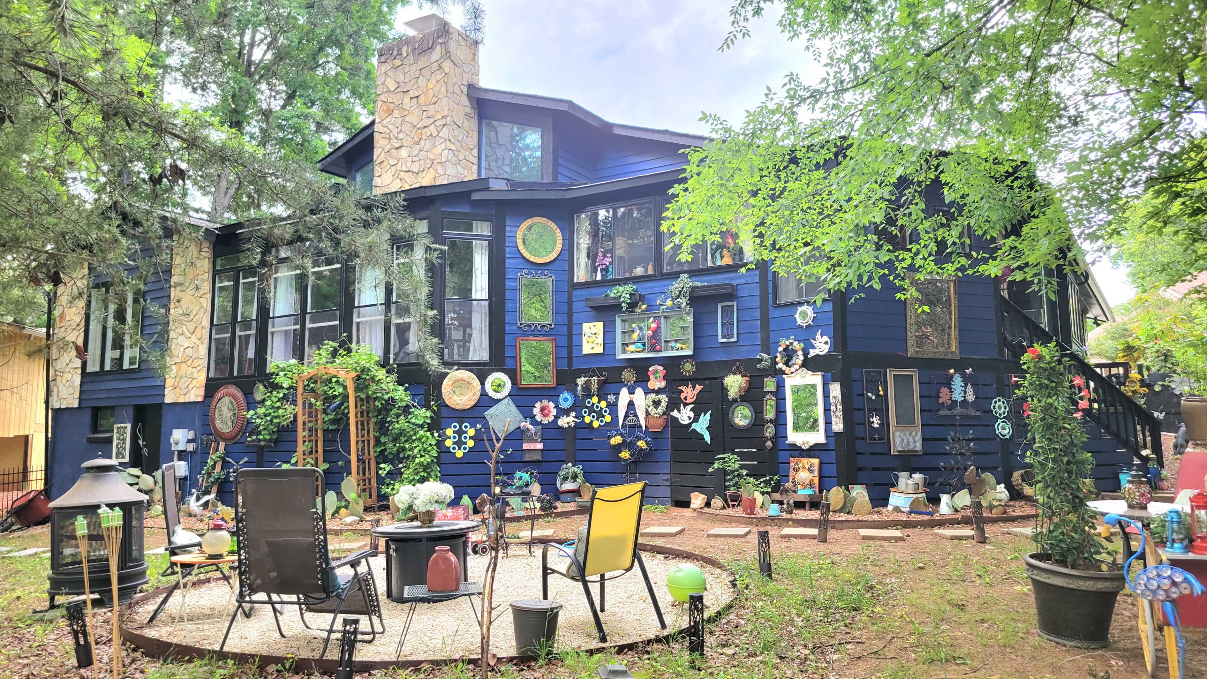 A large, dark blue house features numerous colorful decorations and art pieces on its exterior, surrounded by a circular stone patio with chairs and a fire pit.