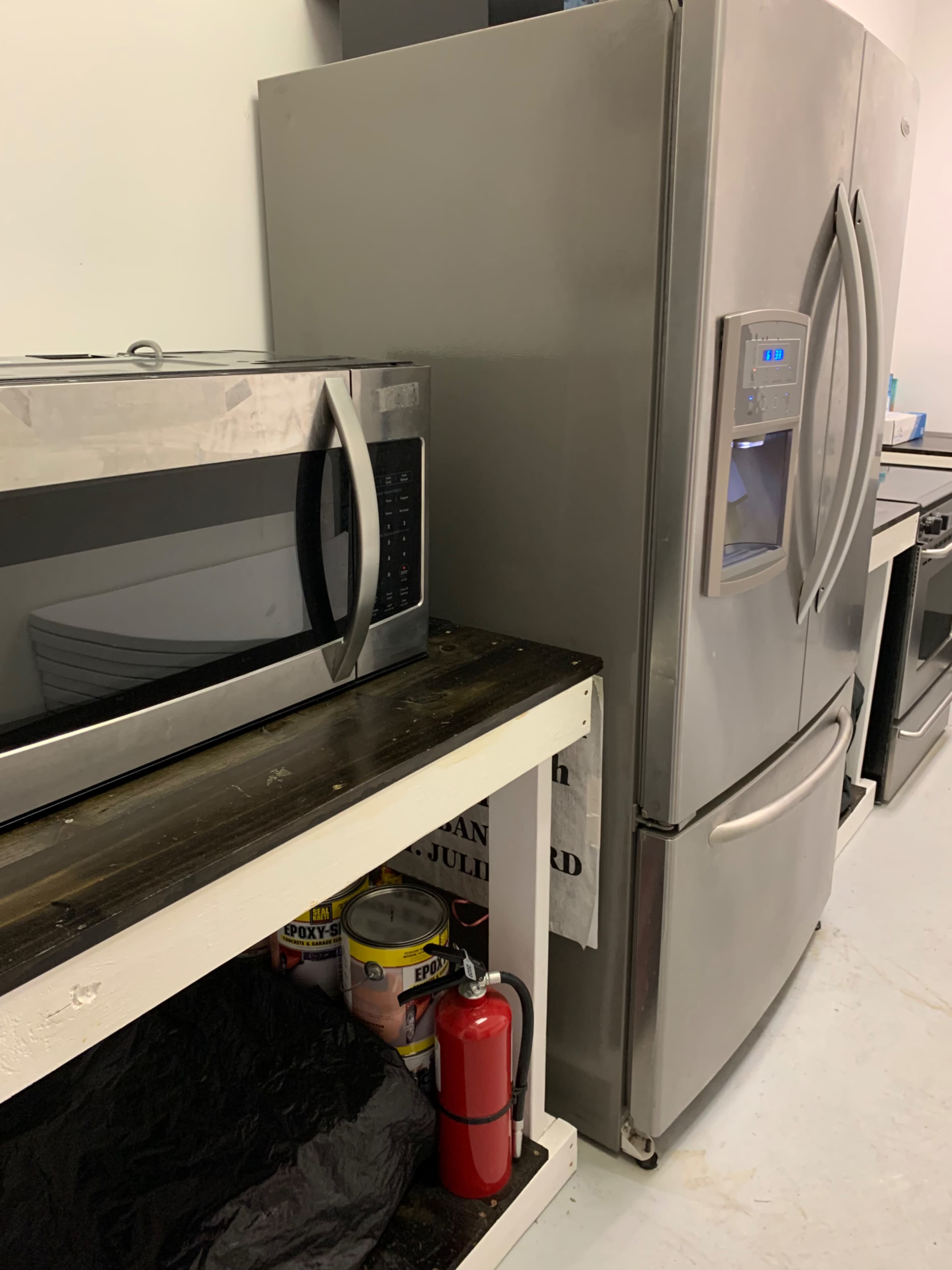 A silver refrigerator is positioned next to a microwave on a wooden shelf, with a fire extinguisher and some canned goods visible underneath.