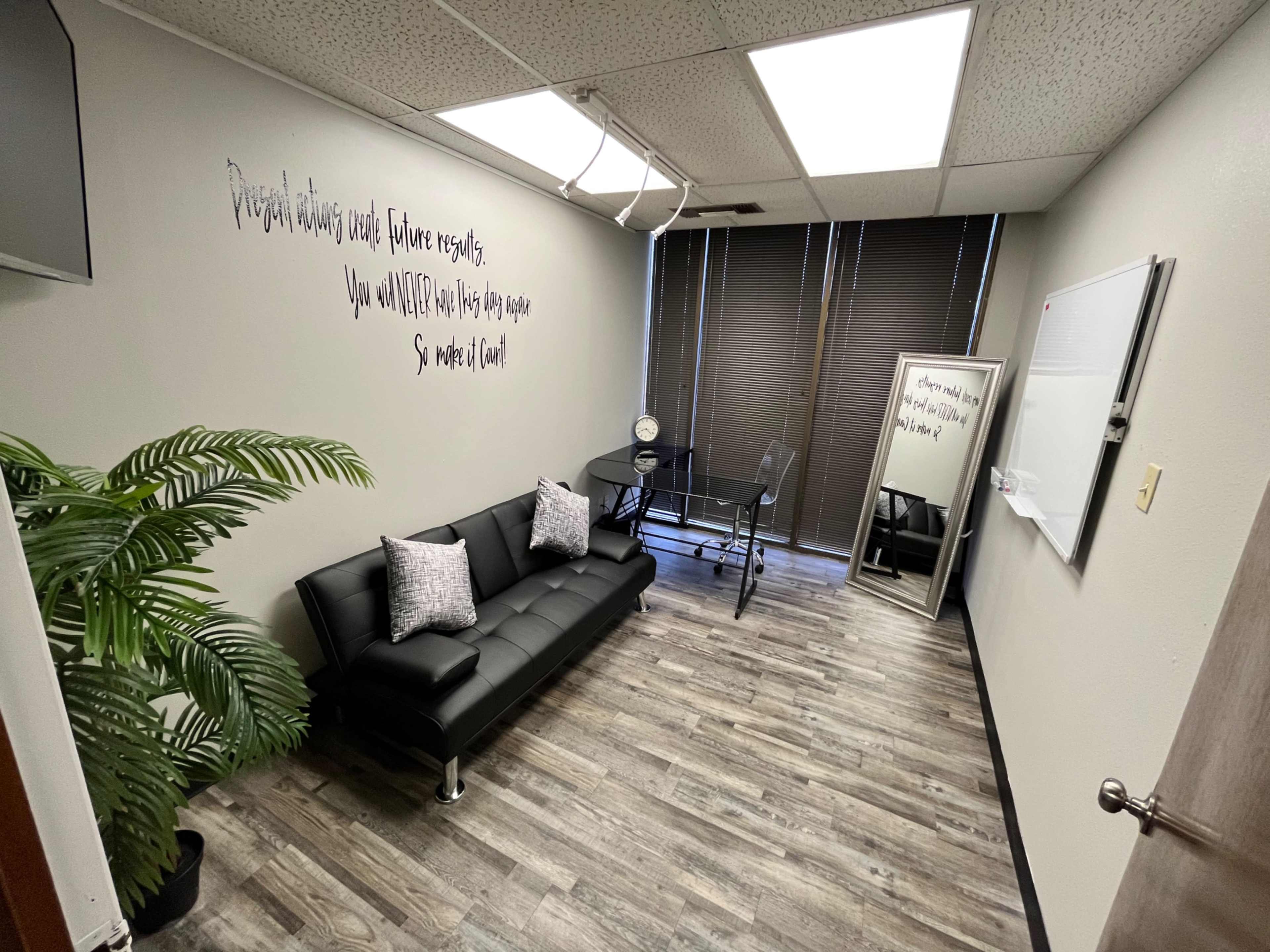 A small office room featuring a black sofa, a glass table with chairs, a large mirror, and a decorative plant against the wall.