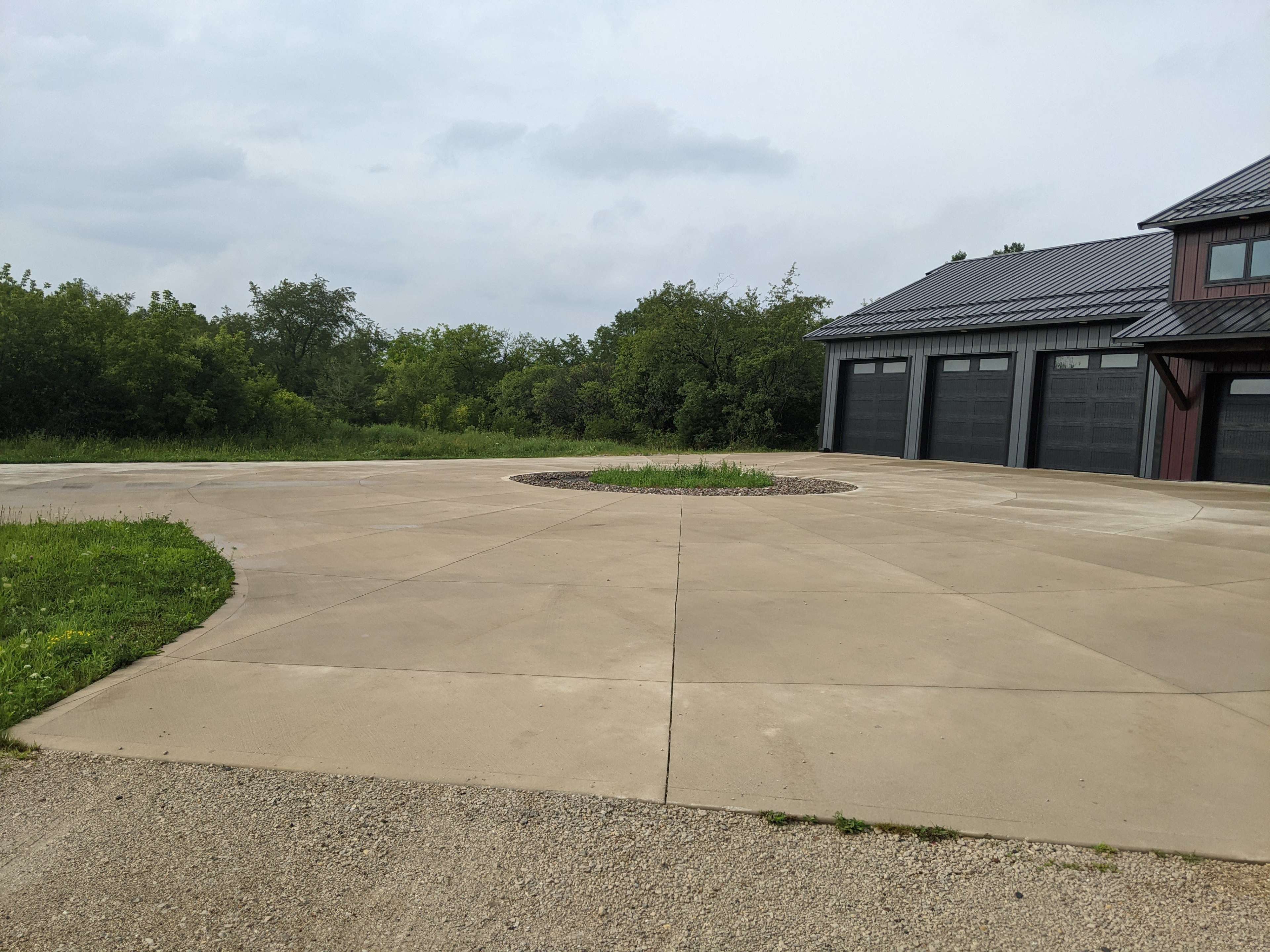 A wide, empty concrete driveway with a circular garden feature in the center, surrounded by greenery and a building with multiple garage doors.