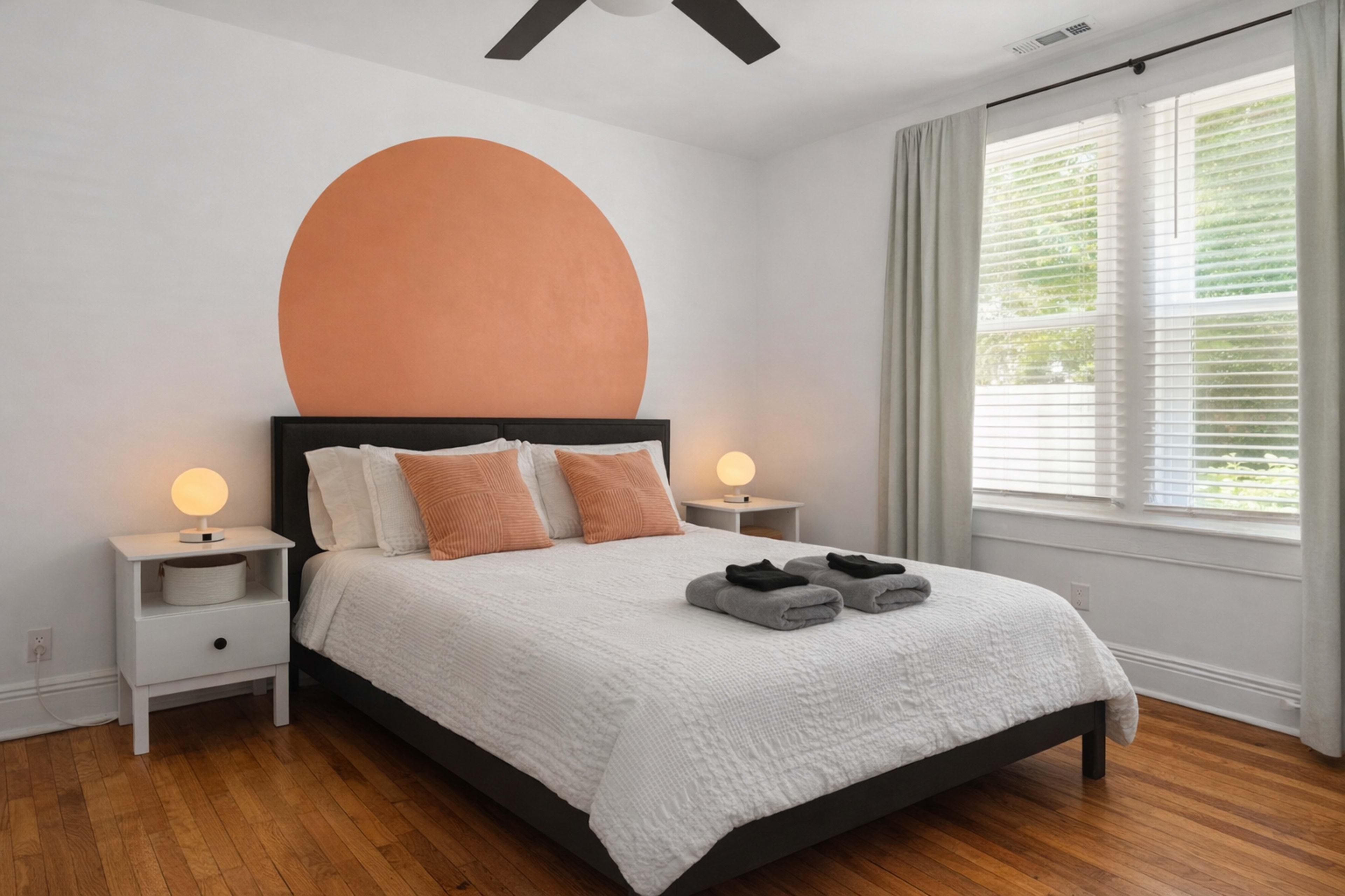 A minimalist bedroom features a bed with white bedding, two side tables with lamps, and a large circular orange accent on the wall behind the bed.