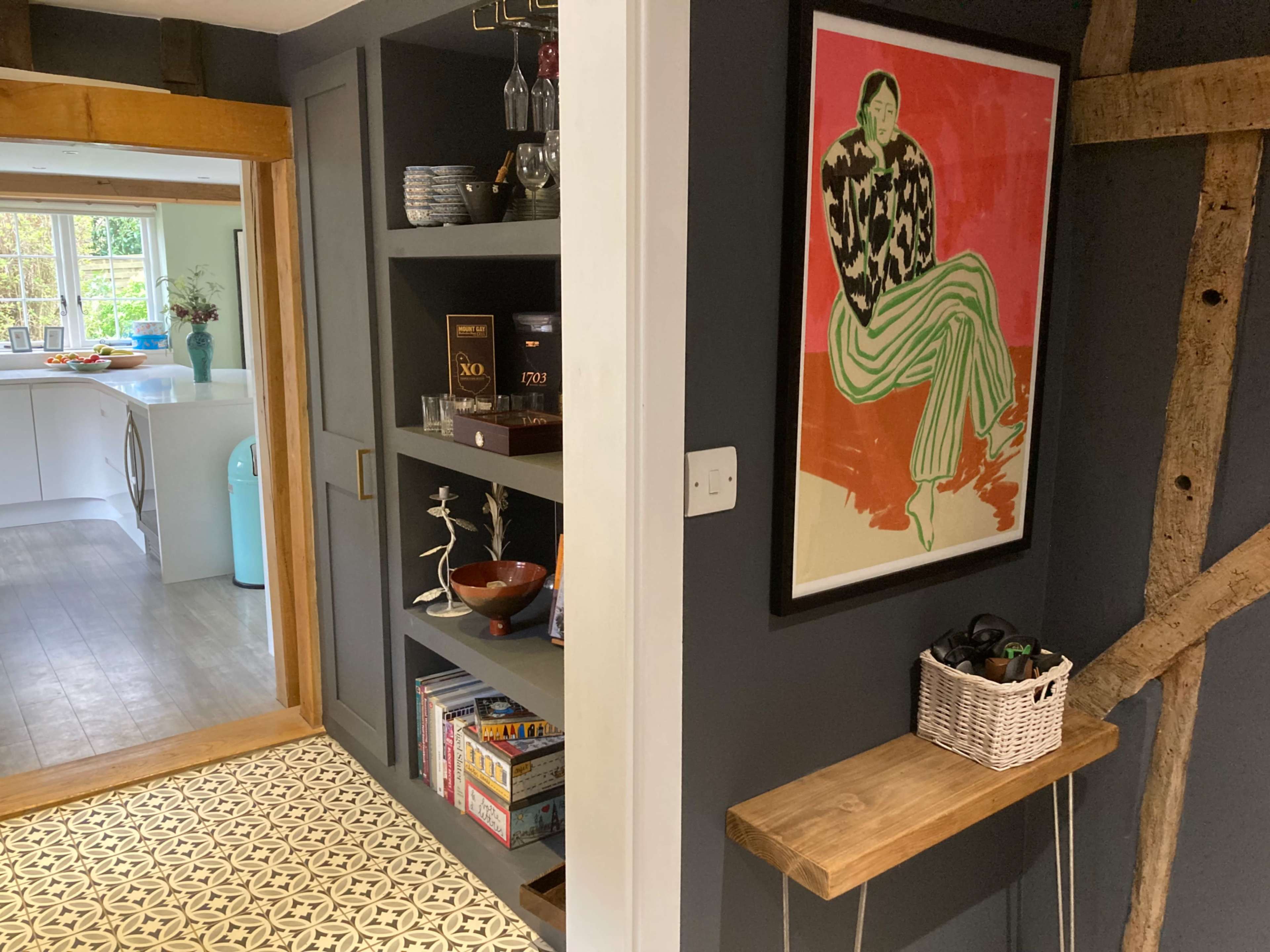 A corridor leading to a kitchen, featuring a wall with a colorful framed artwork, shelves filled with books and decorative items, and a wooden shelf with a basket.