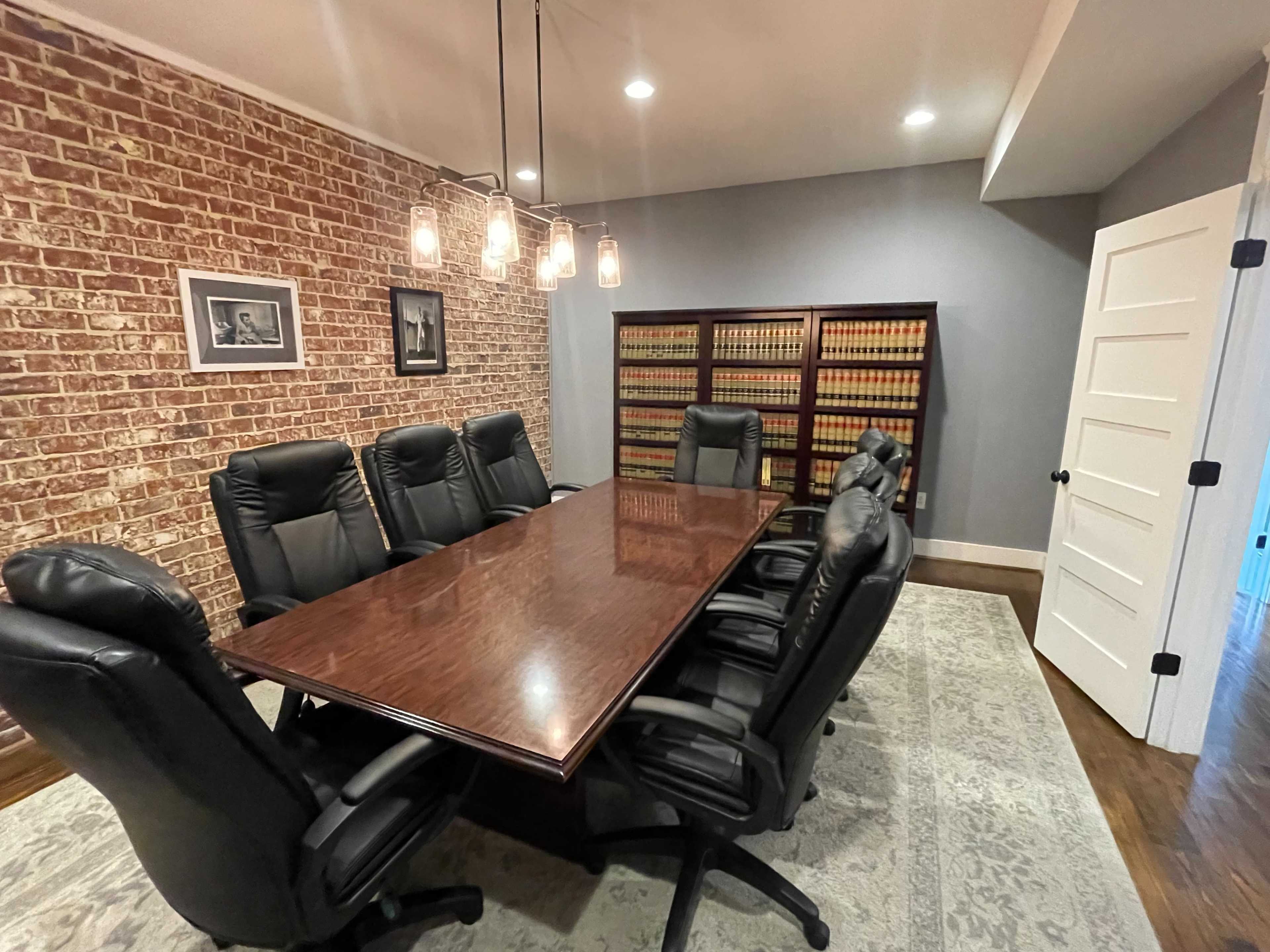 A conference room with a large wooden table surrounded by black leather chairs, brick walls, and a bookshelf filled with legal books.