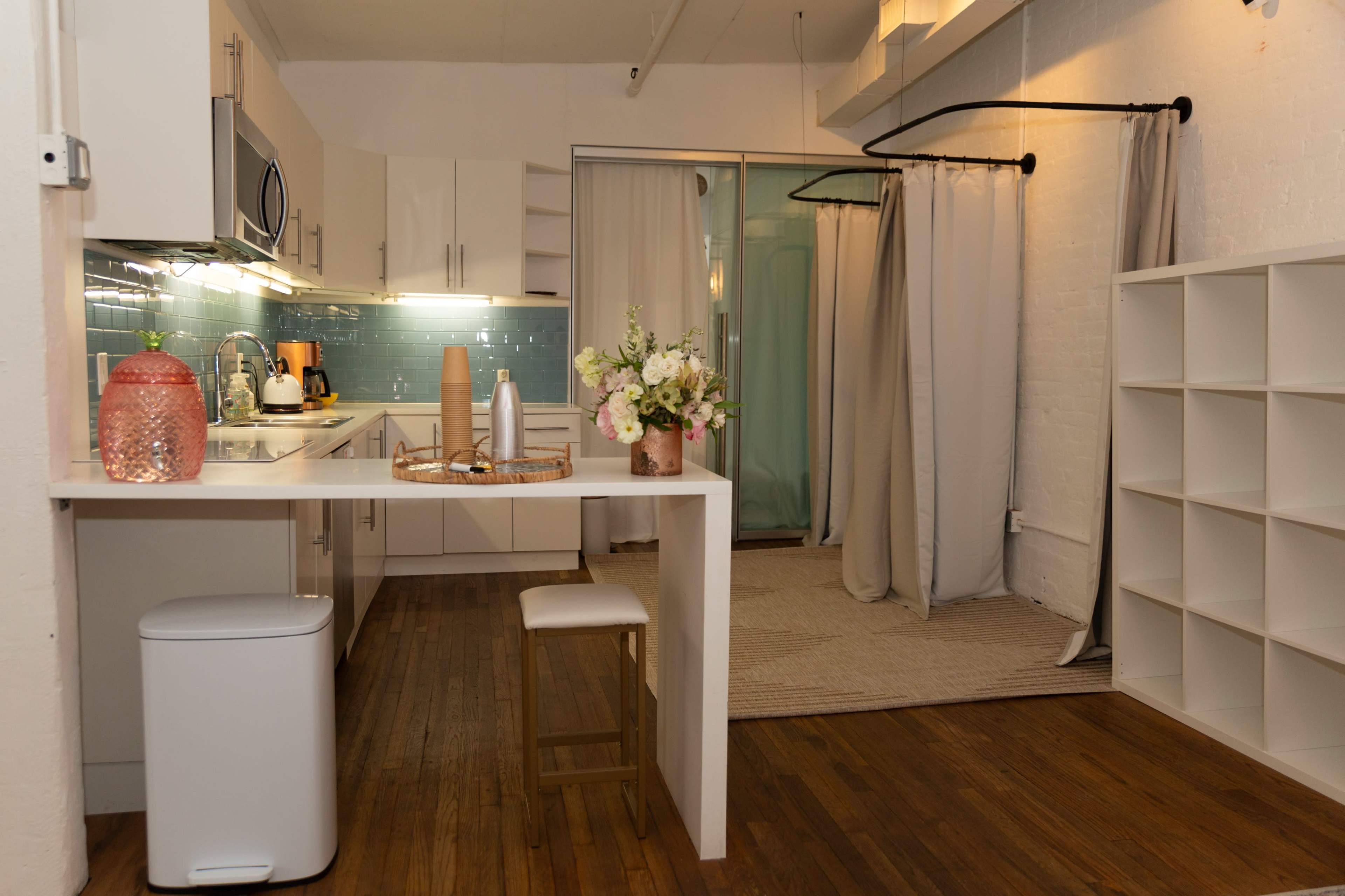 The image shows a modern kitchen area with white cabinetry, a blue tile backsplash, and a connected living space featuring a curtain divider and a light-colored rug.