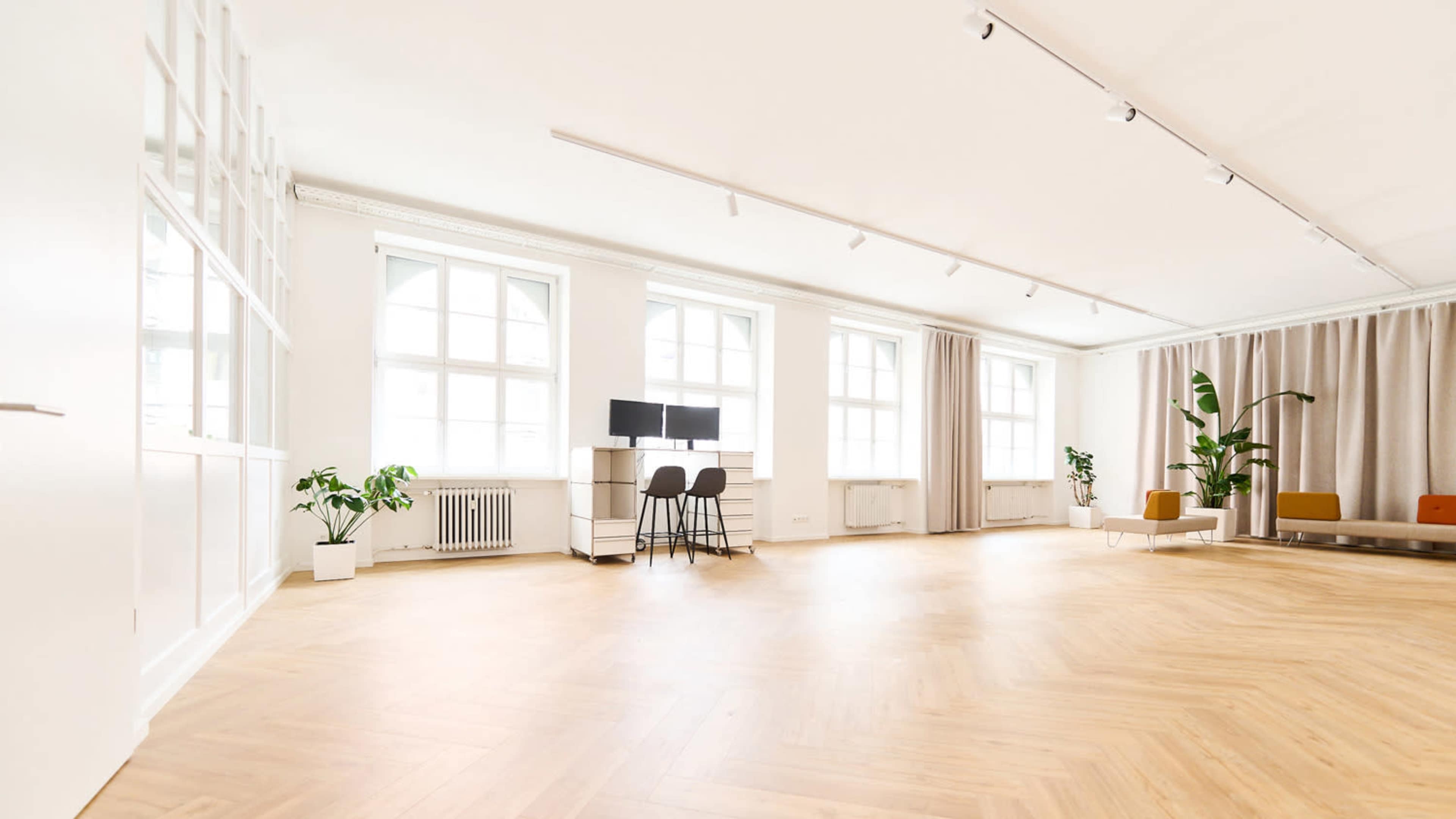 The image shows a spacious, well-lit room with large windows, wooden flooring, and minimalistic furniture including a seating area and a workspace.