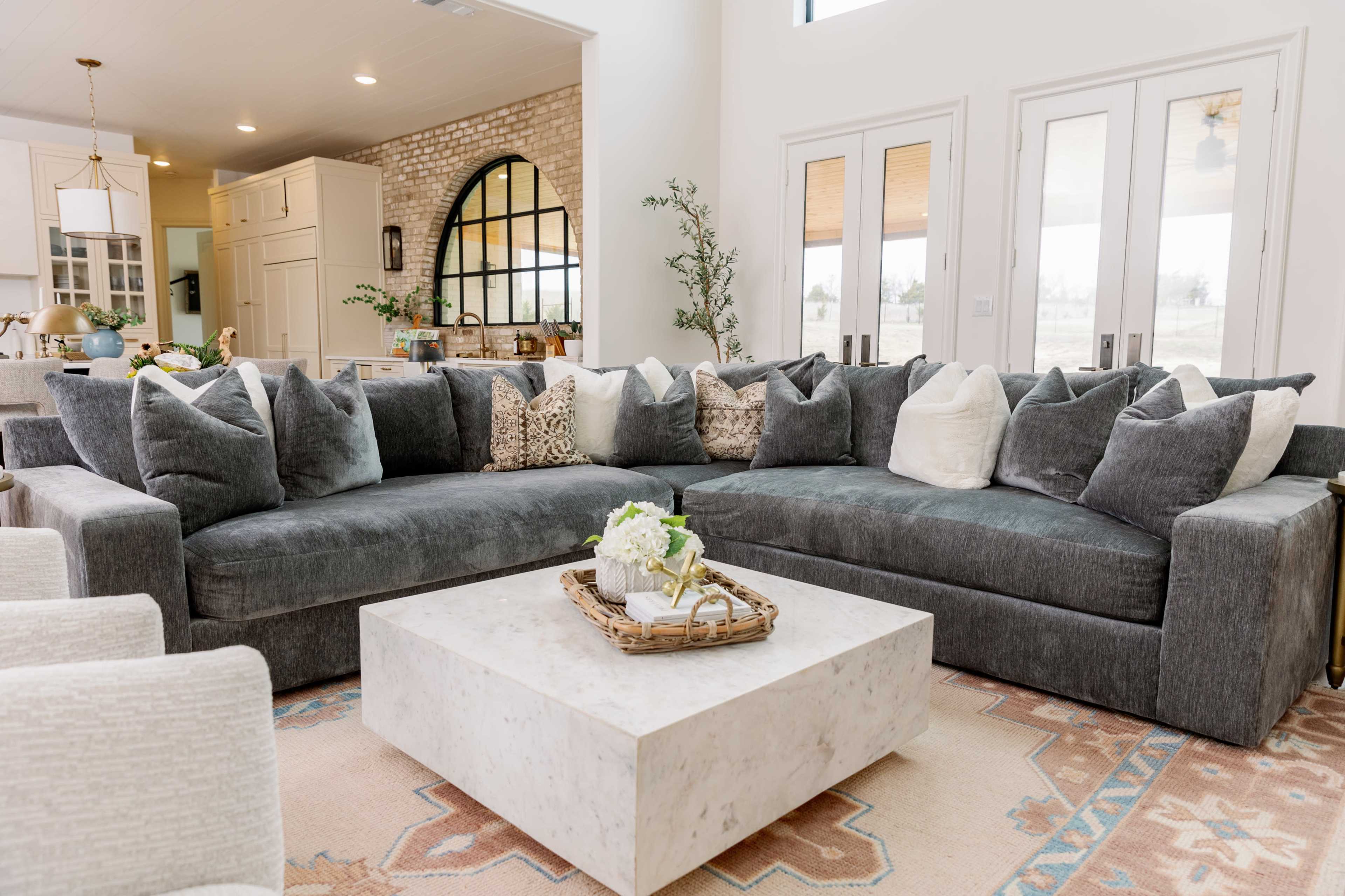The image shows a large, gray sectional sofa adorned with multiple patterned and solid throw pillows, positioned near a marble coffee table on a decorative rug in a bright, open living space.