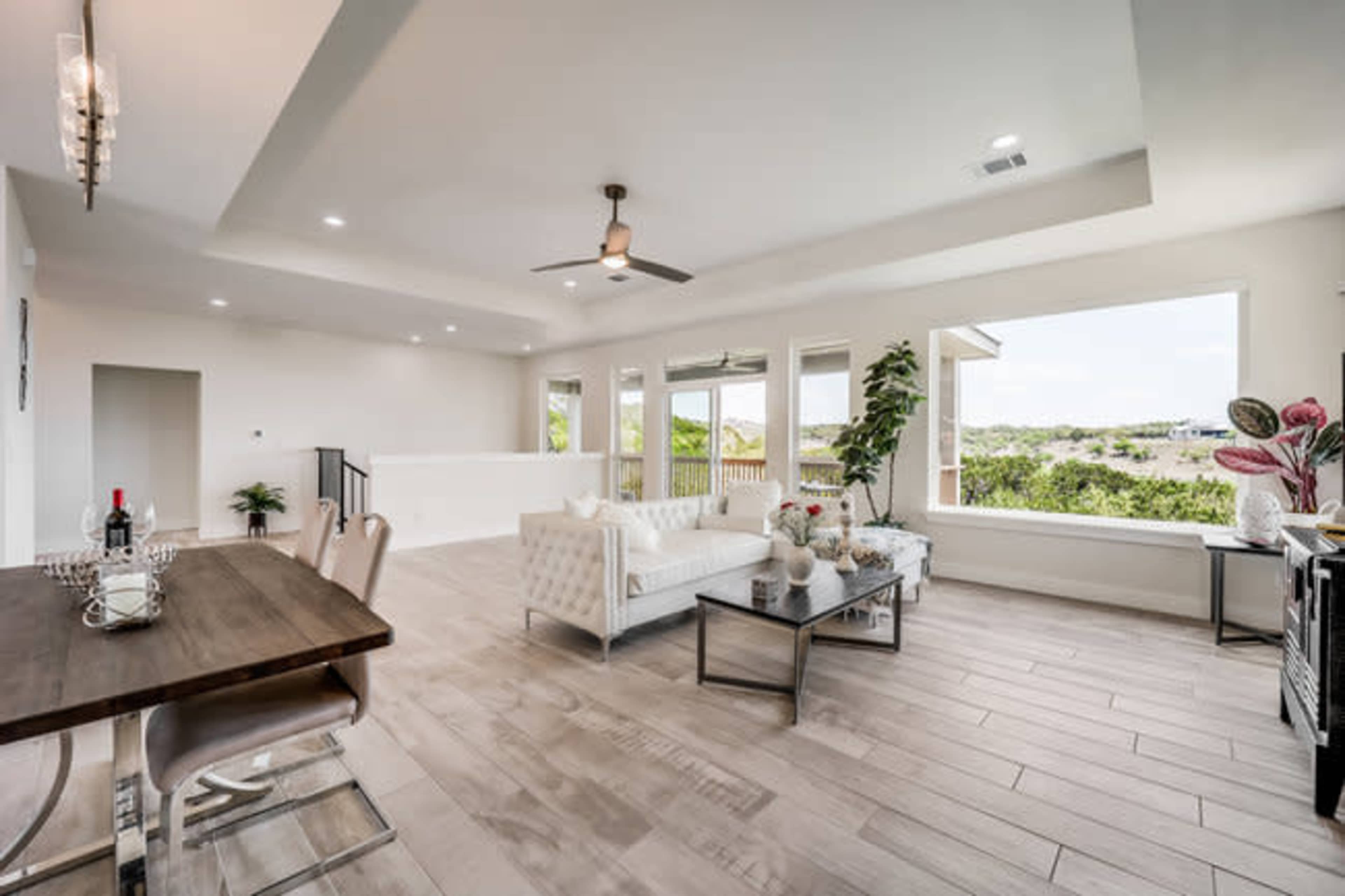 The image shows a modern, open-concept living area with a dining table, a white sectional sofa, large windows offering a view of the outside, and neutral-colored walls and flooring.