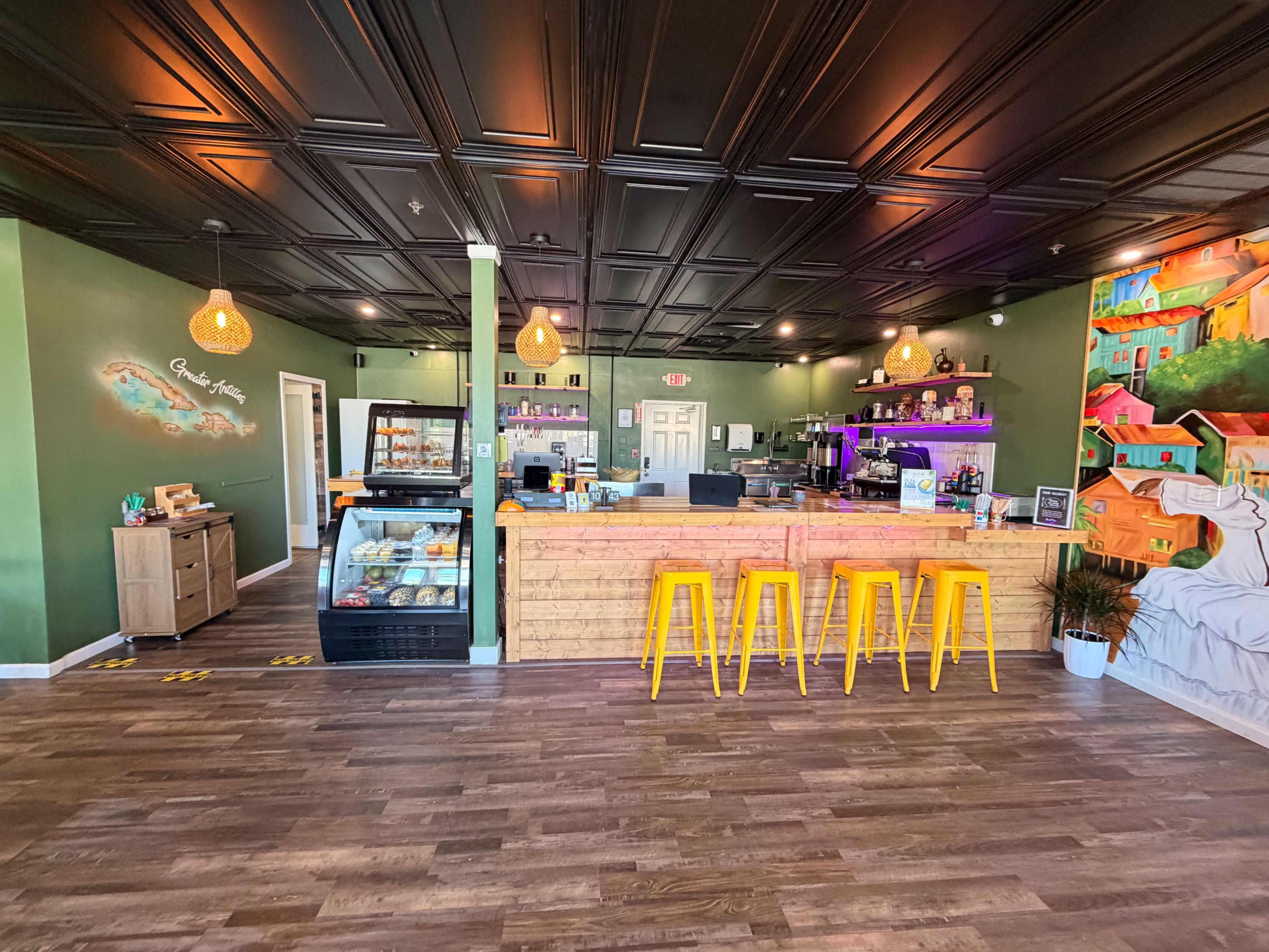 The image shows a brightly colored café interior with a wooden counter, yellow barstools, and a mural on the wall.