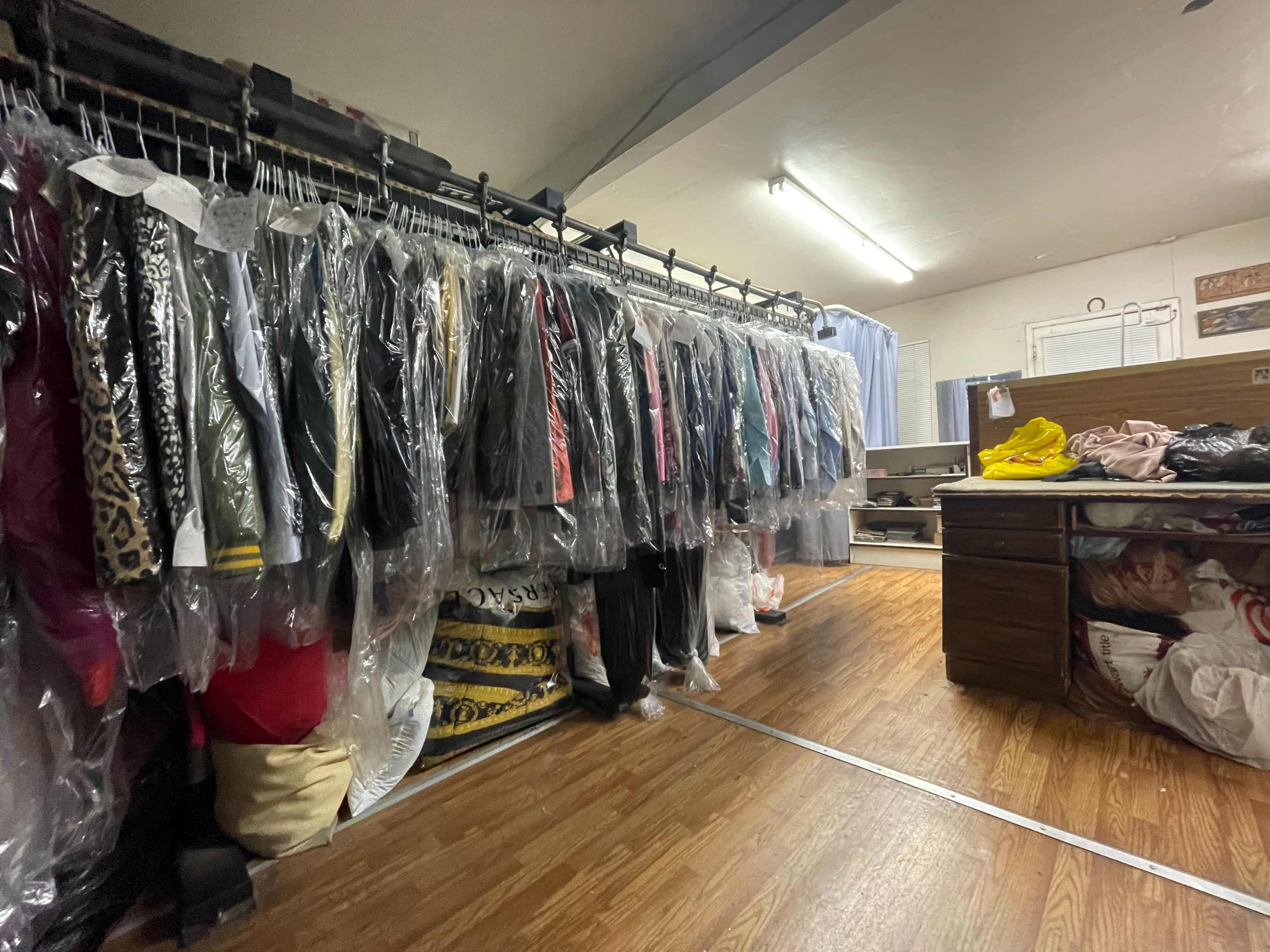 A room with racks of clothing covered in plastic and a cluttered workspace with a bed and various textiles.