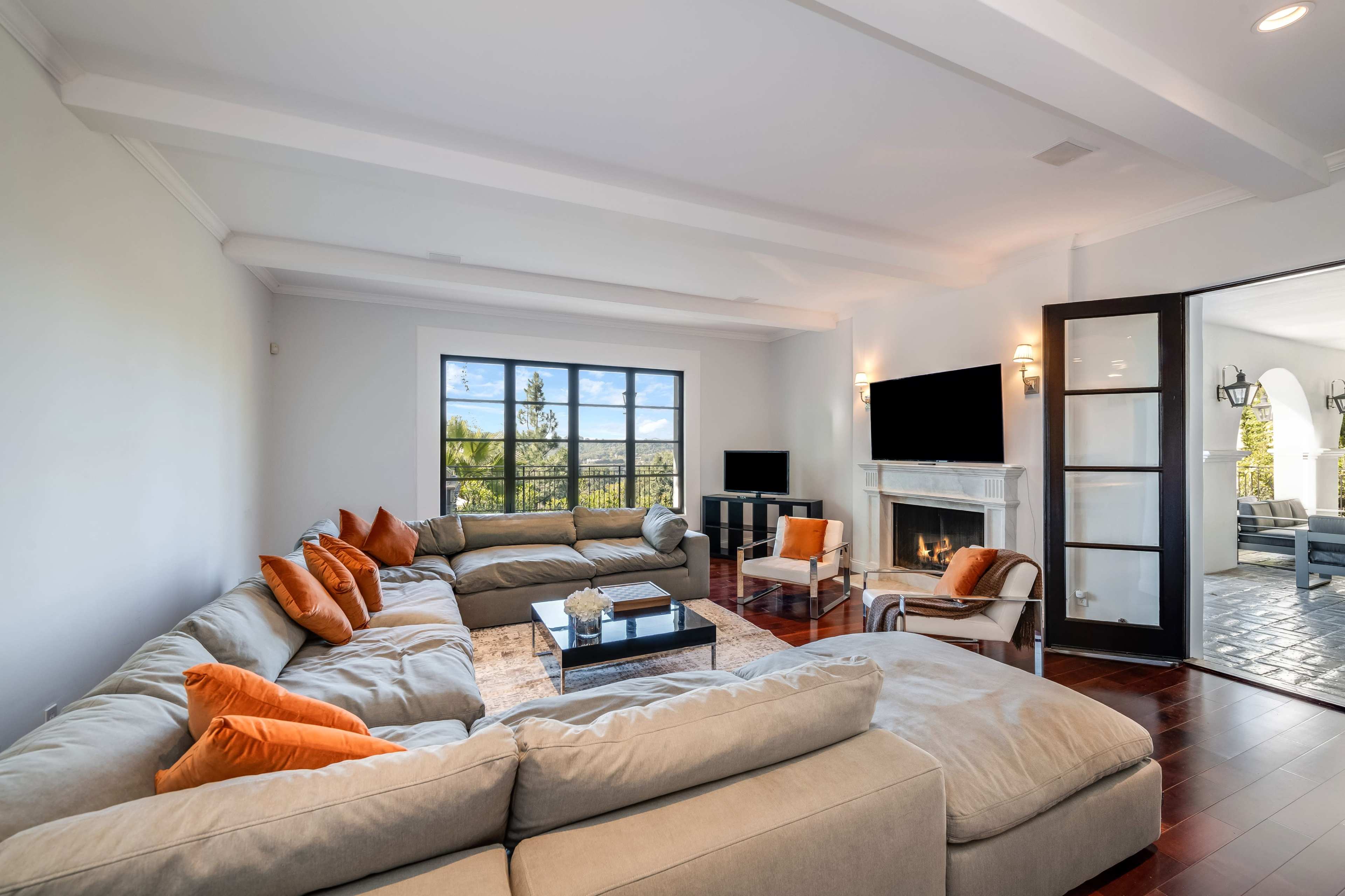 The image shows a spacious living room featuring a large sectional sofa with orange accent pillows, a coffee table, a fireplace, and a window overlooking an outdoor view.