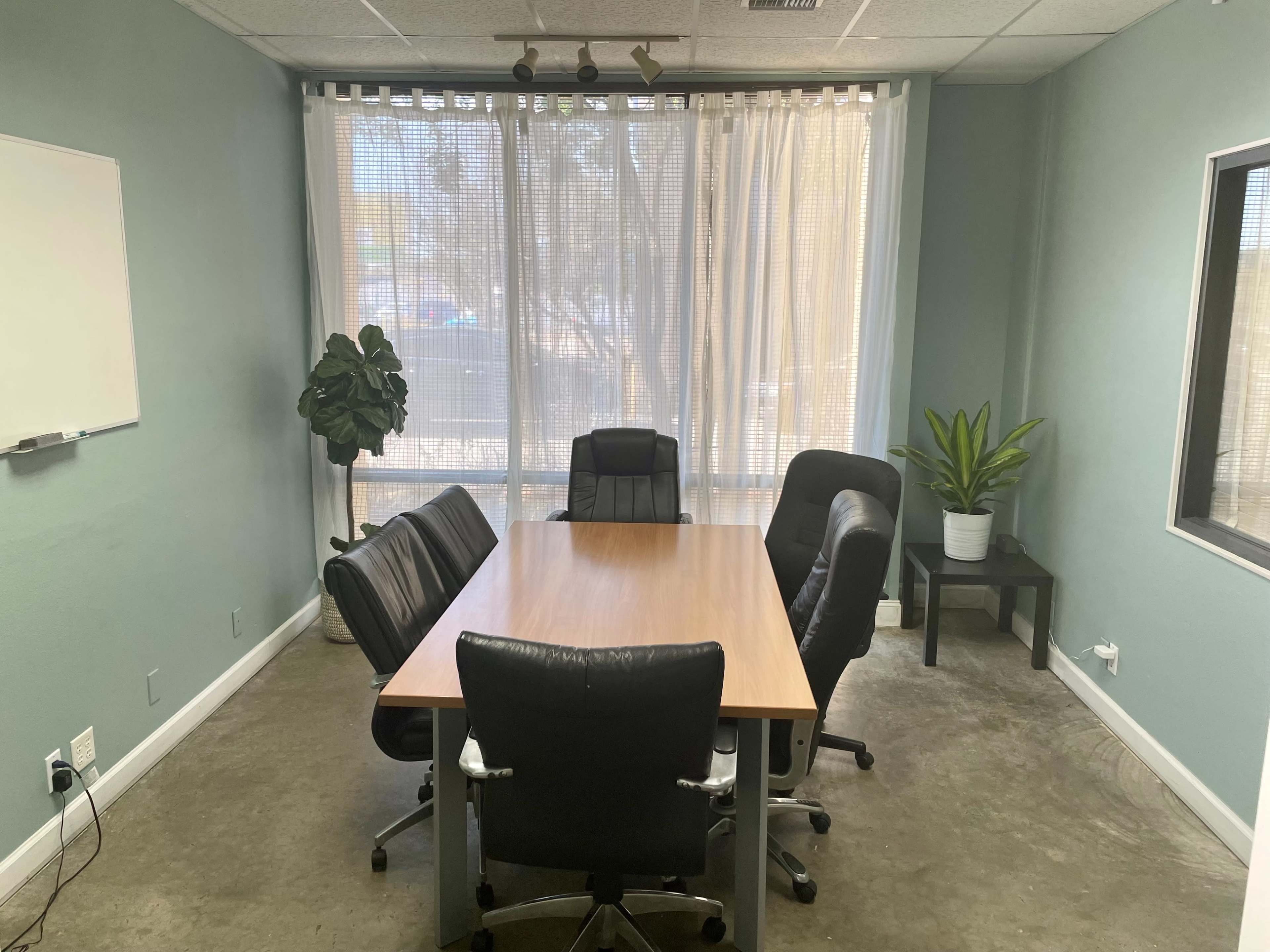 A conference room features a rectangular wooden table surrounded by five black office chairs, with a large window covered by sheer curtains and two potted plants in the corners.