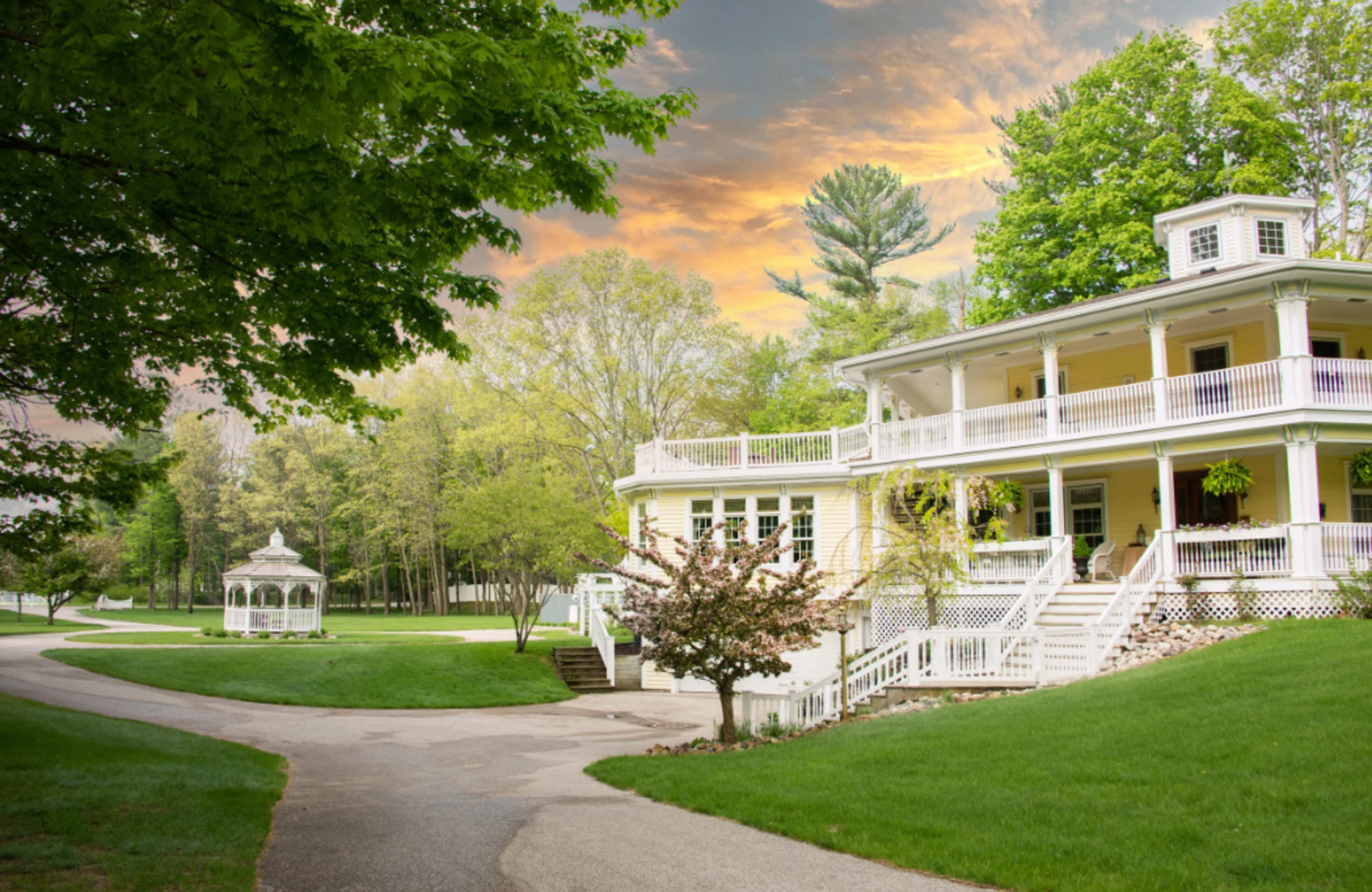 A large, two-story yellow house with a wraparound porch and a white gazebo sits amidst lush greenery under a colorful sky at sunset.