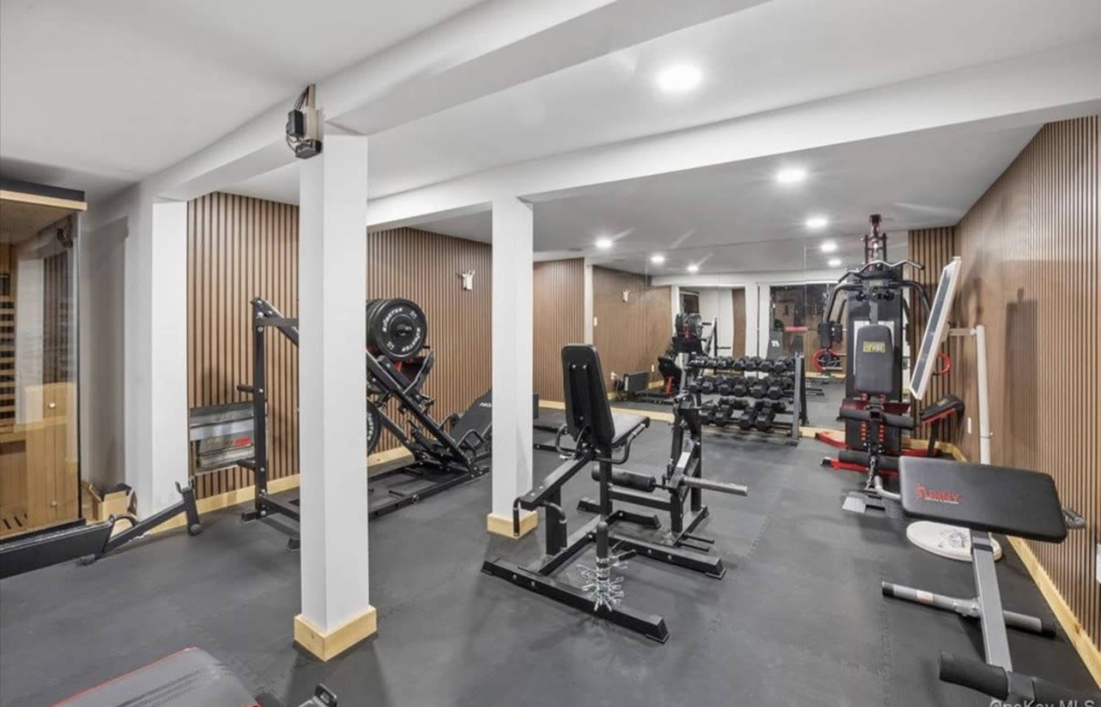 The image shows a modern home gym equipped with various fitness machines, weights, and exercise equipment in a well-lit space.