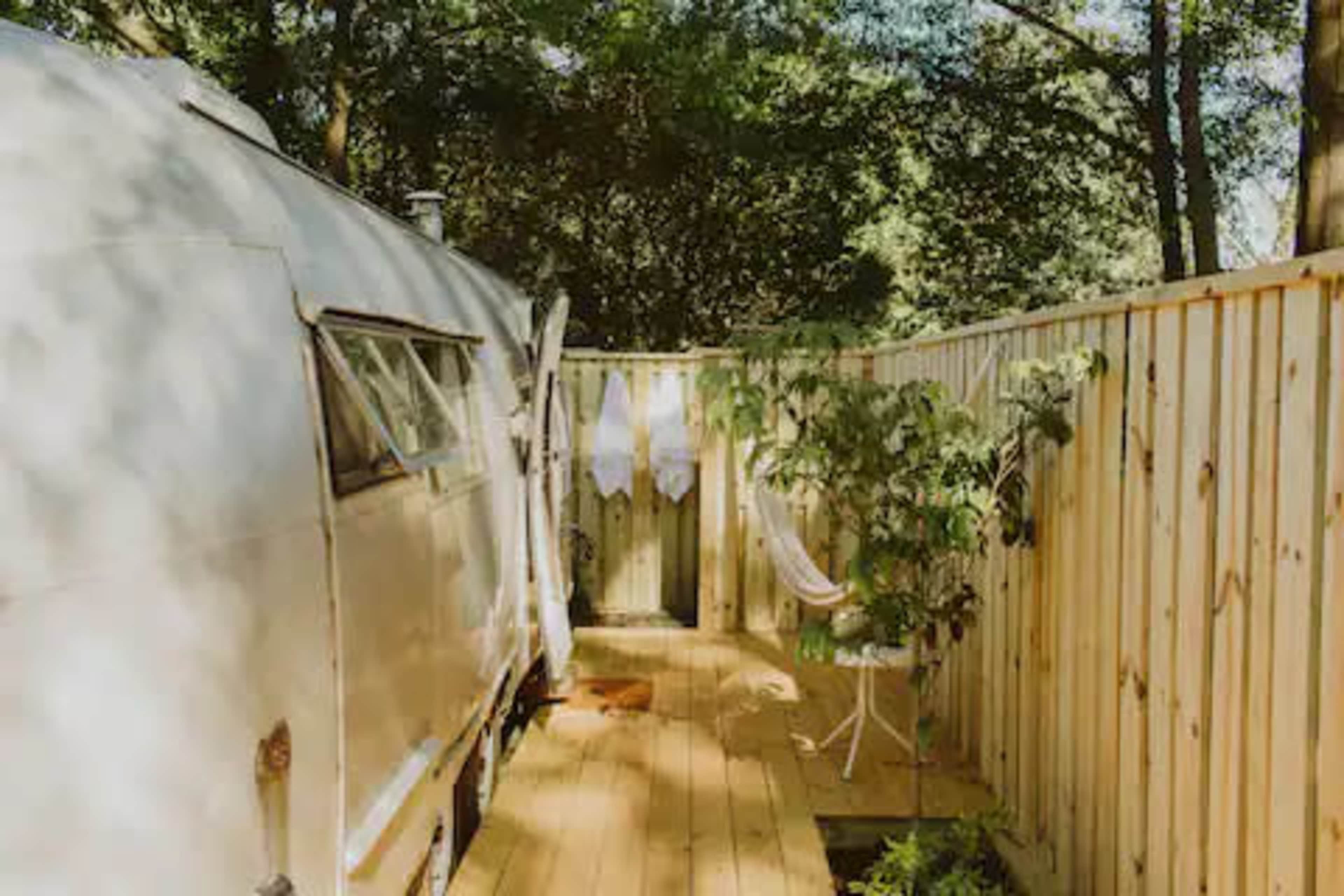 A camper is parked beside a wooden deck, surrounded by a fenced area with potted plants and a hanging chair.