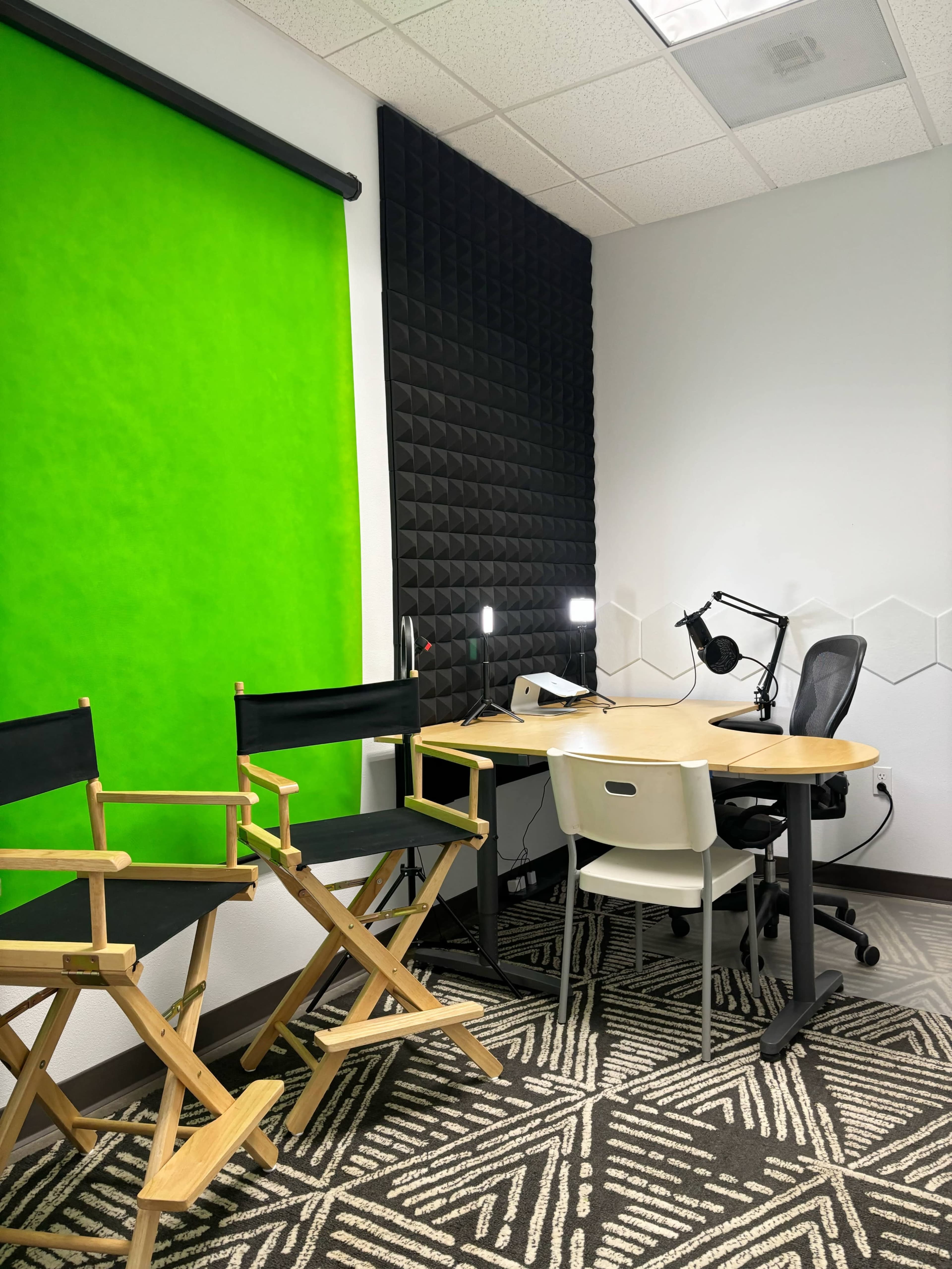 A small studio setup featuring two director's chairs, a green backdrop, a desk with a microphone attached, and acoustic paneling on one wall.