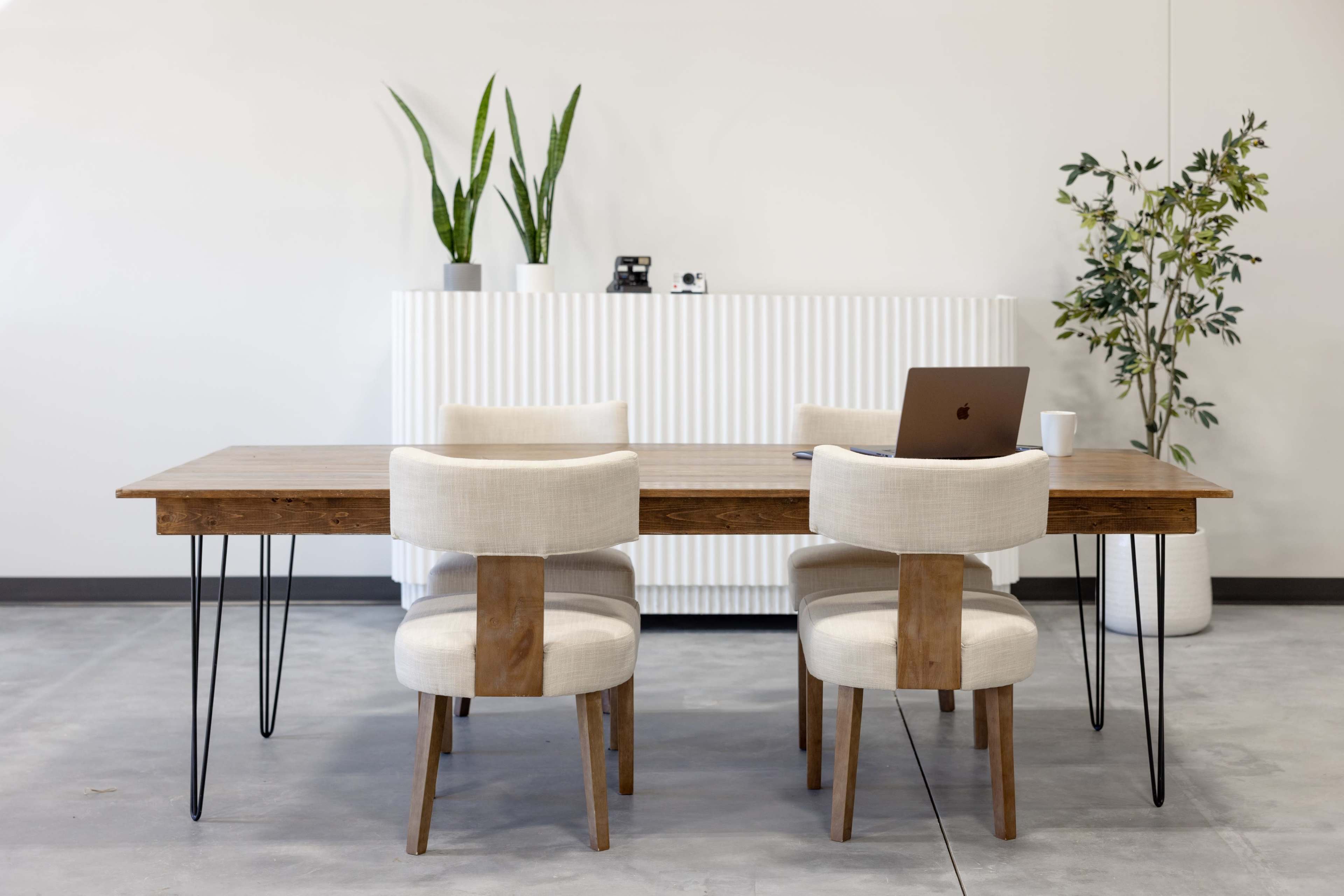 A modern dining table is set with four chairs, accompanied by a laptop and a white cup on the table, while a potted plant and a tall vase stand in the background.