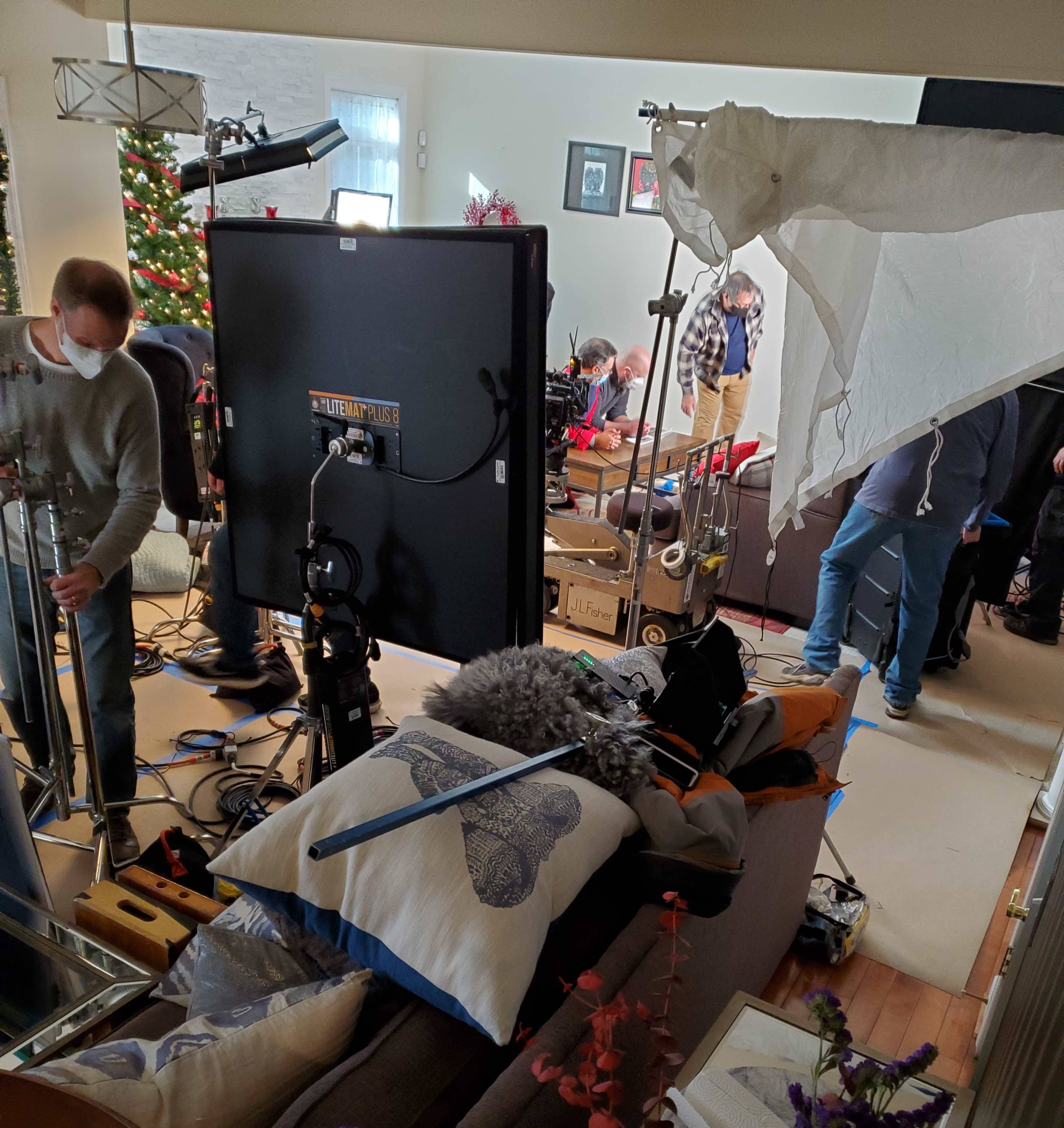 A film crew sets up equipment in a living room decorated for the holidays, featuring a Christmas tree and various filming gear scattered around.