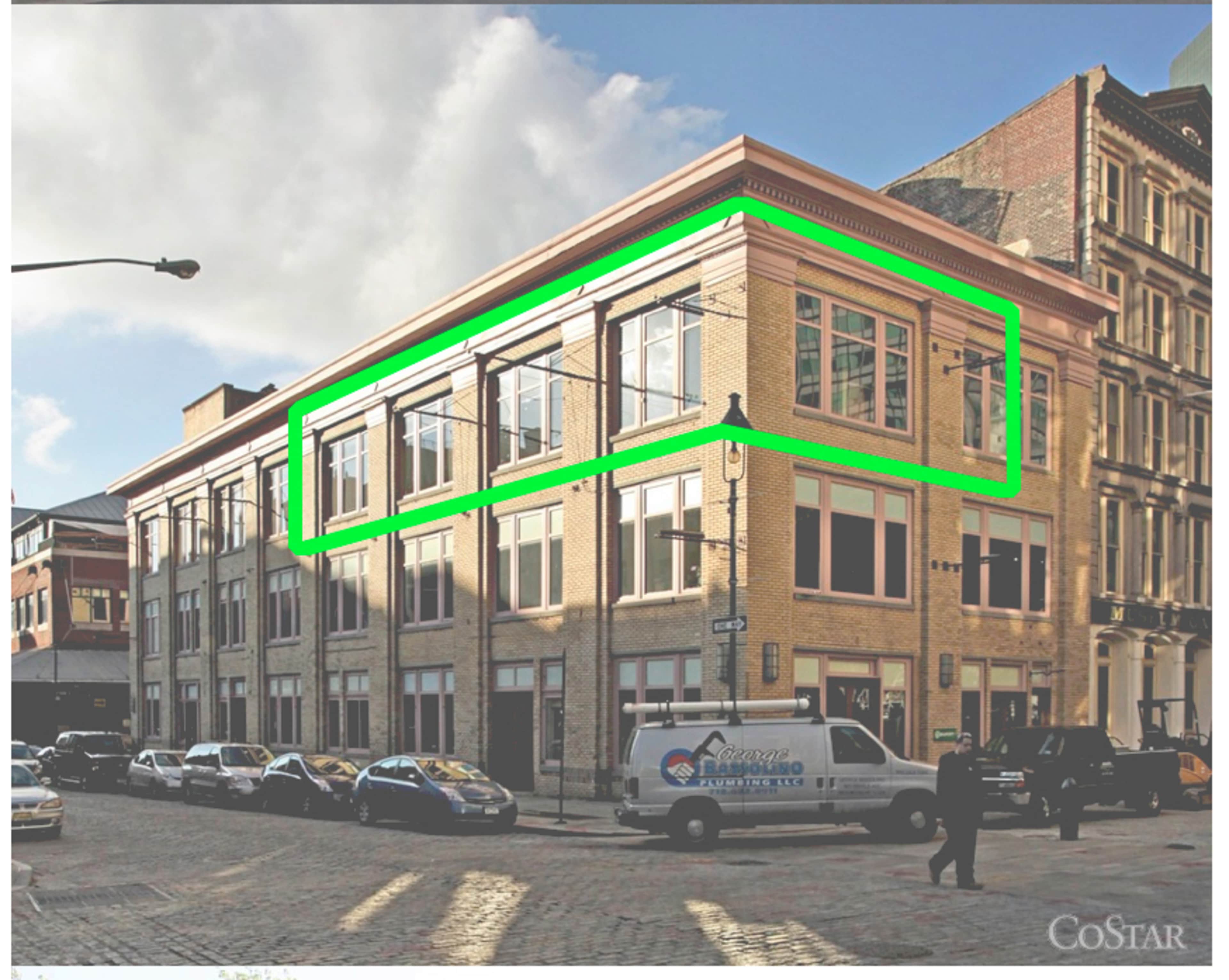 A brick building with large windows on the upper level, located on a city street with parked vehicles.