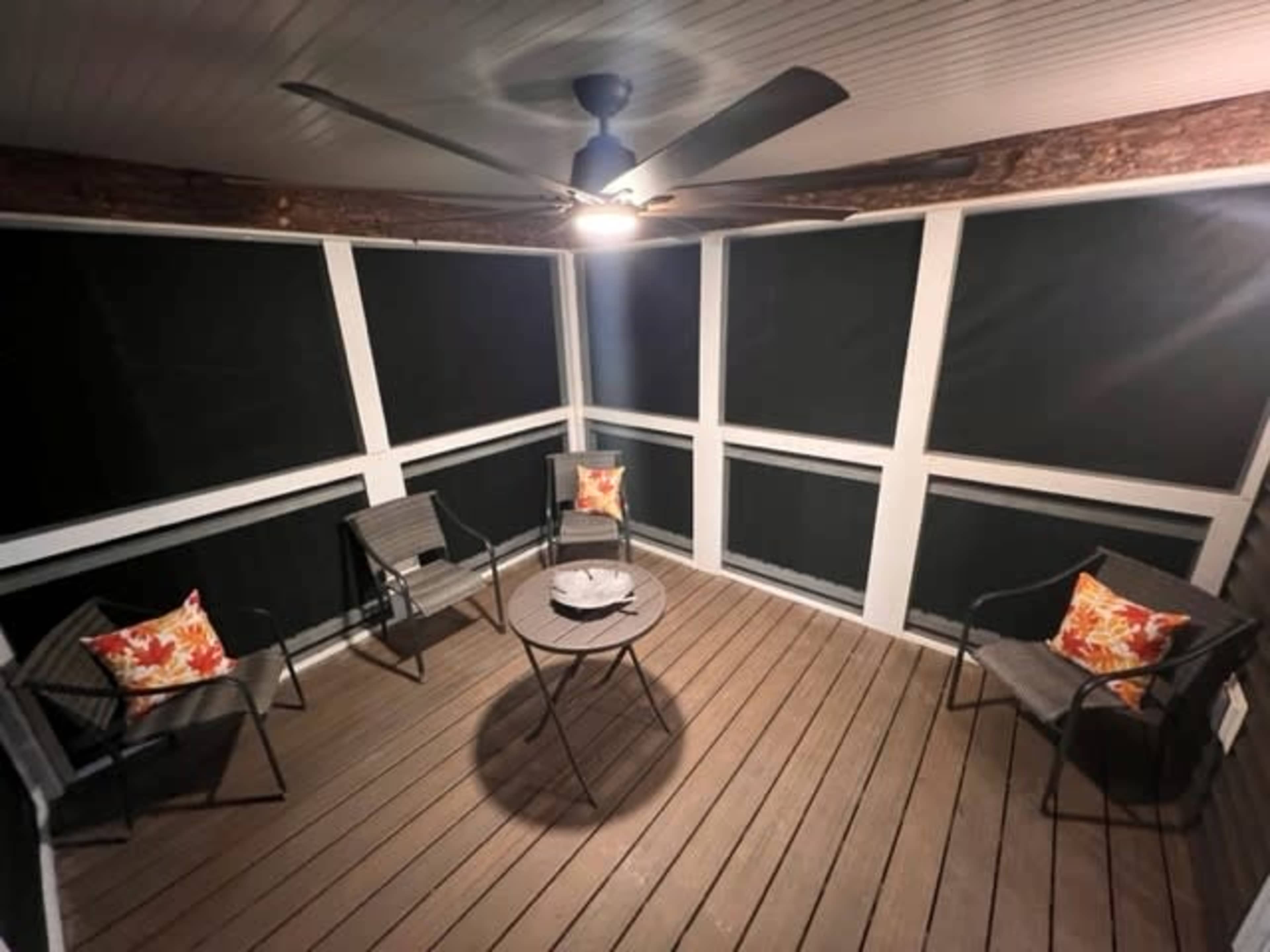 A screened-in porch features a circular table and four chairs, with decorative pillows, under a ceiling fan.