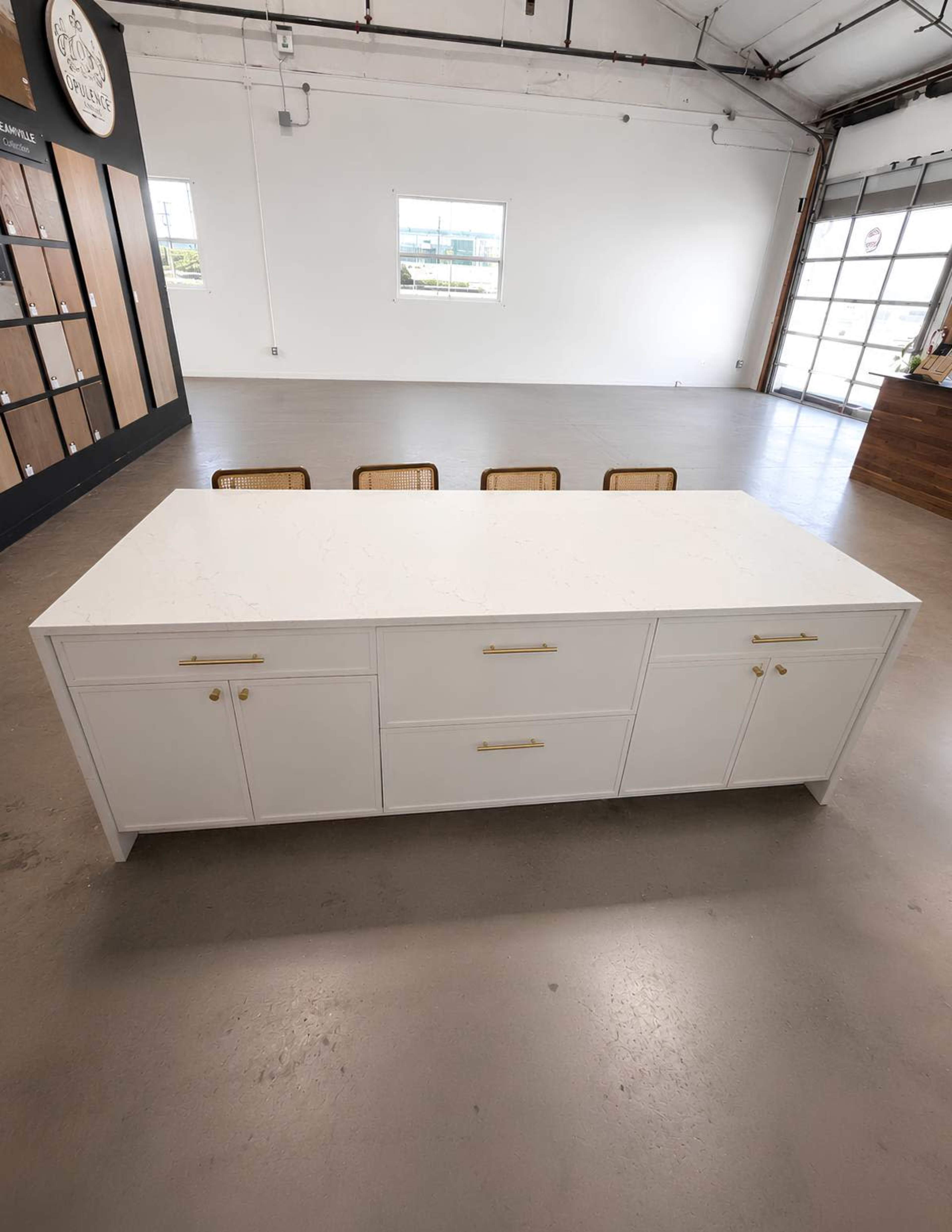 A white kitchen island with gold hardware commands attention in the center of a spacious, well-lit room, surrounded by wooden chairs facing it.