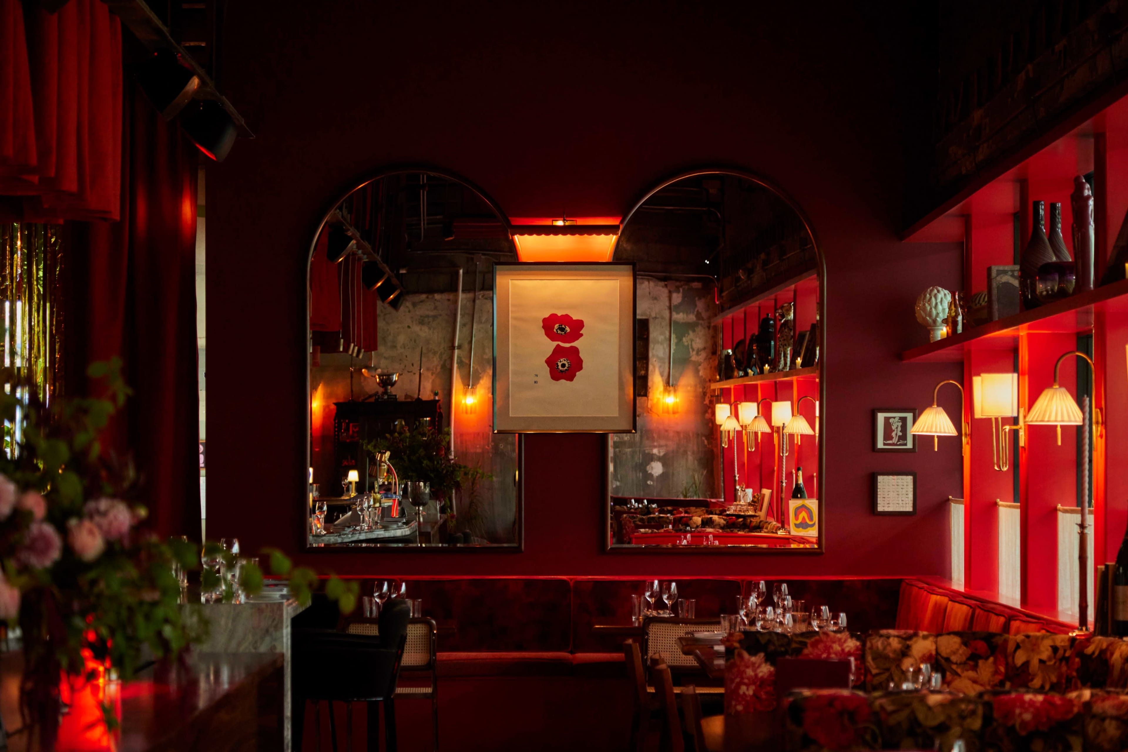 The image shows a dimly lit restaurant interior featuring red walls, decorative mirrors, and a large floral artwork above a bar area.
