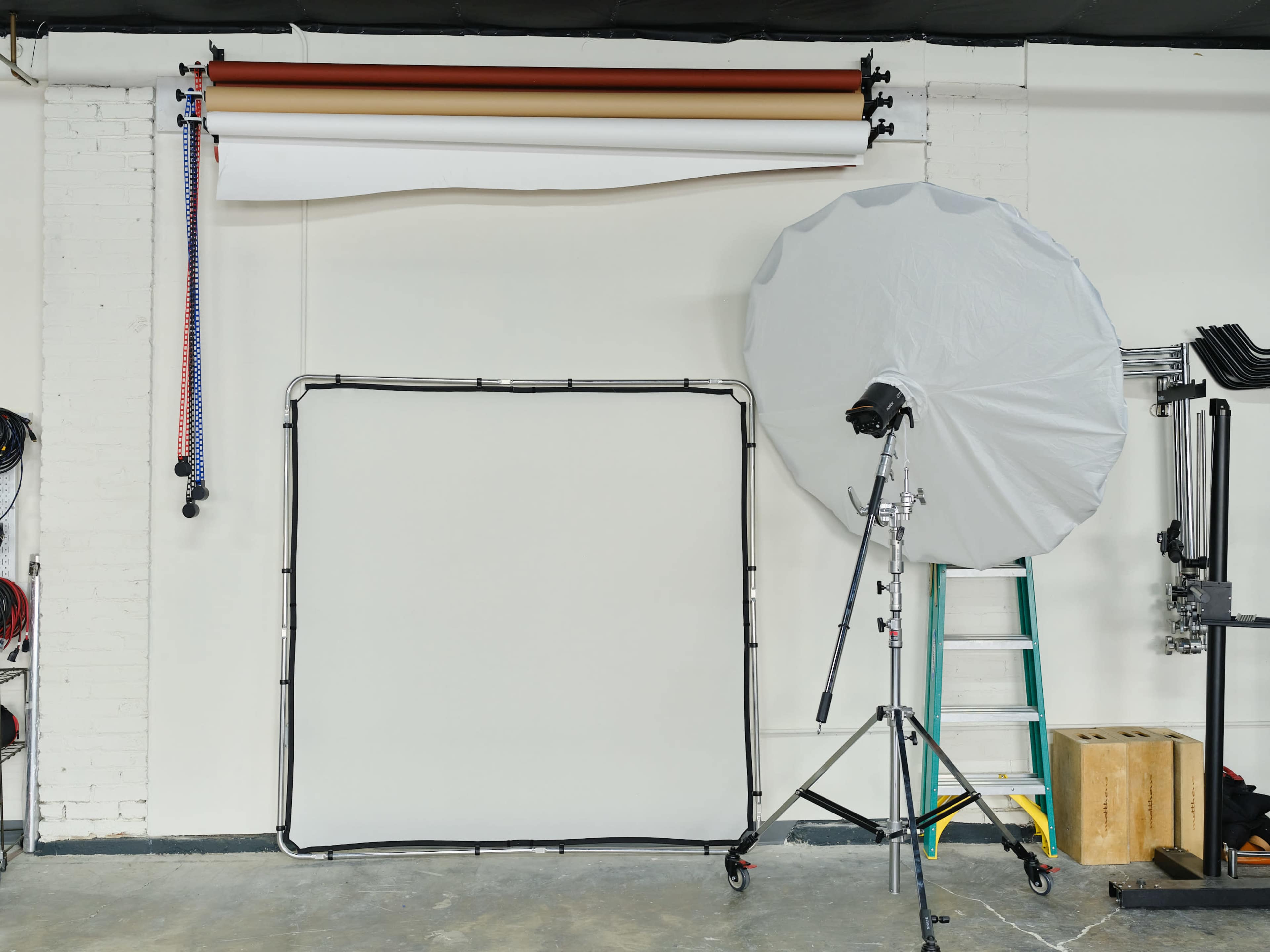 A photography studio setup with a large white backdrop, a lighting umbrella, a folding ladder, and various equipment against a plain wall.