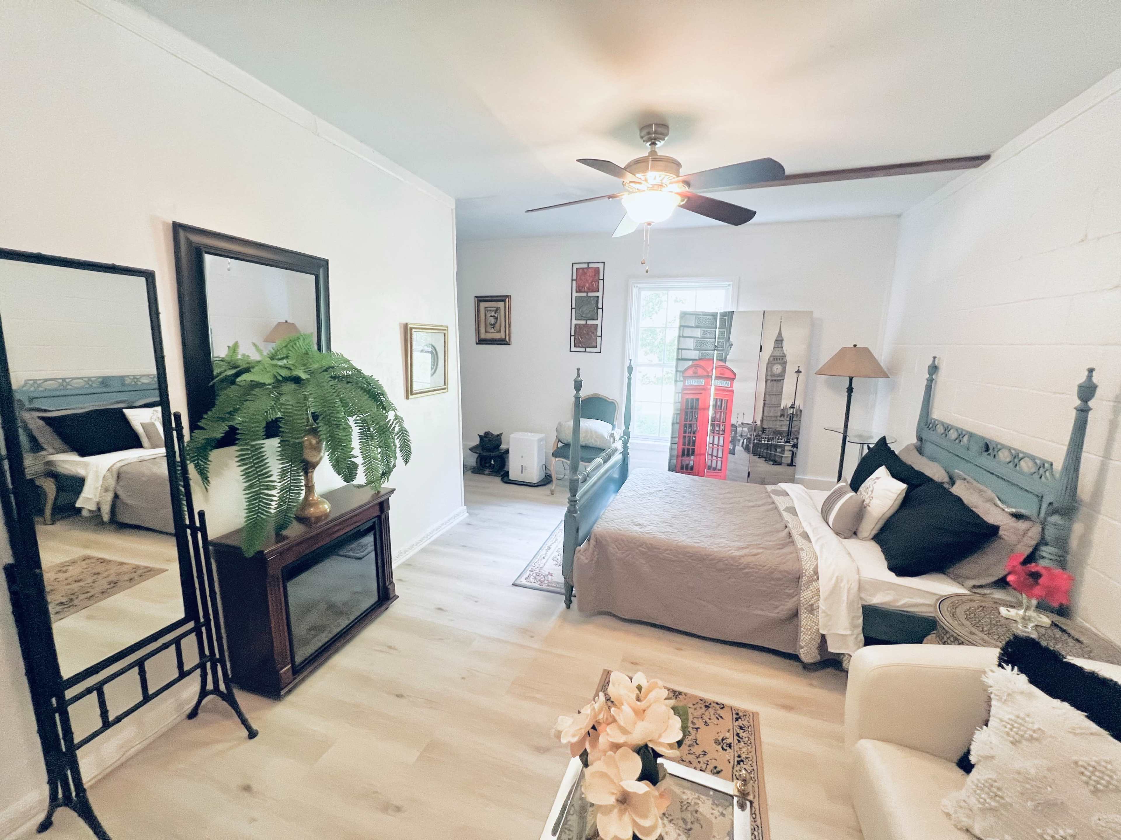 A bright bedroom with two beds, a ceiling fan, and a mirror, alongside a seating area and decorative elements, including a lamp and potted plant.
