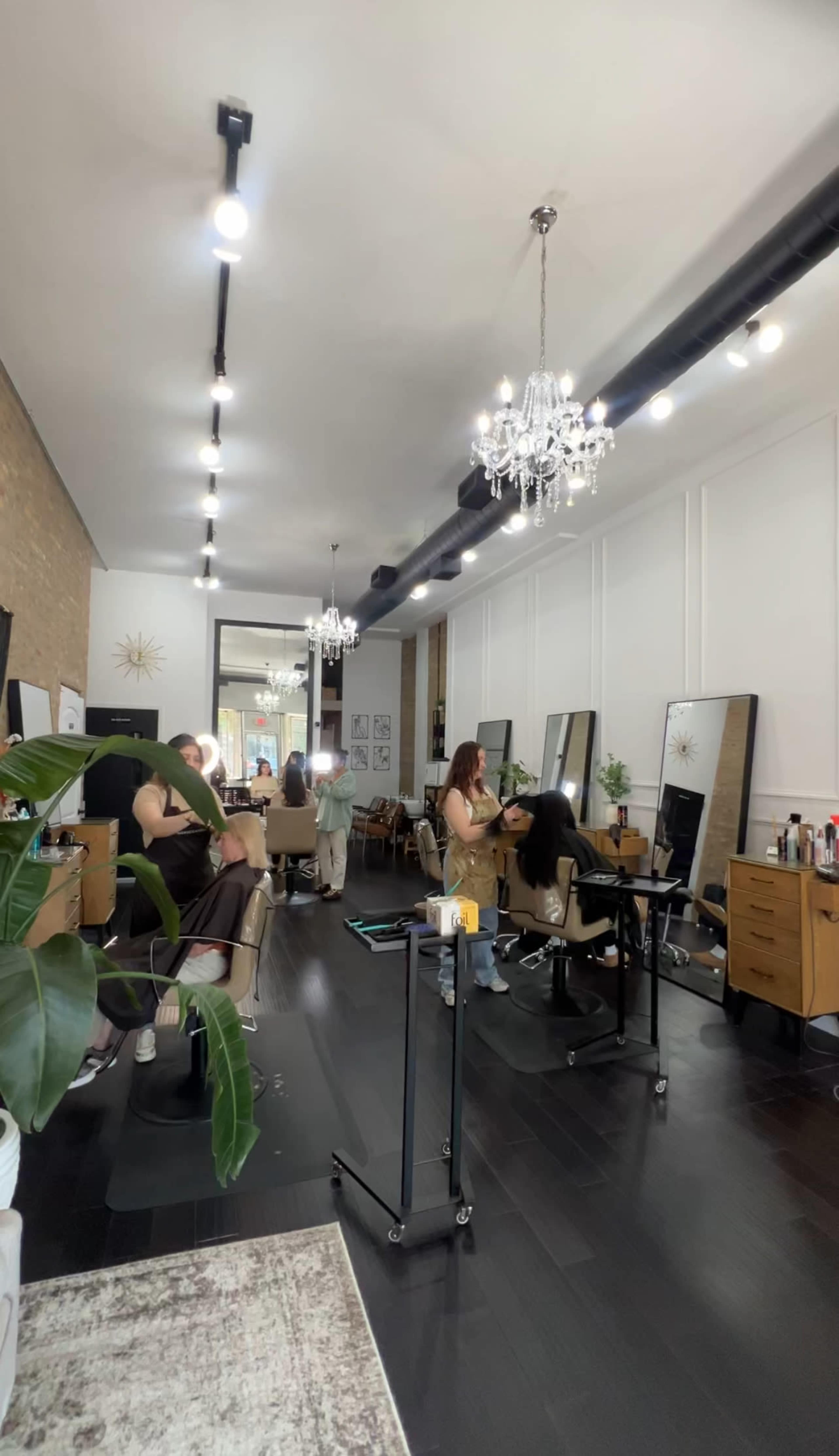 A modern hair salon features several stylist stations with clients and stylists engaged in hair services, illuminated by chandeliers and natural light.