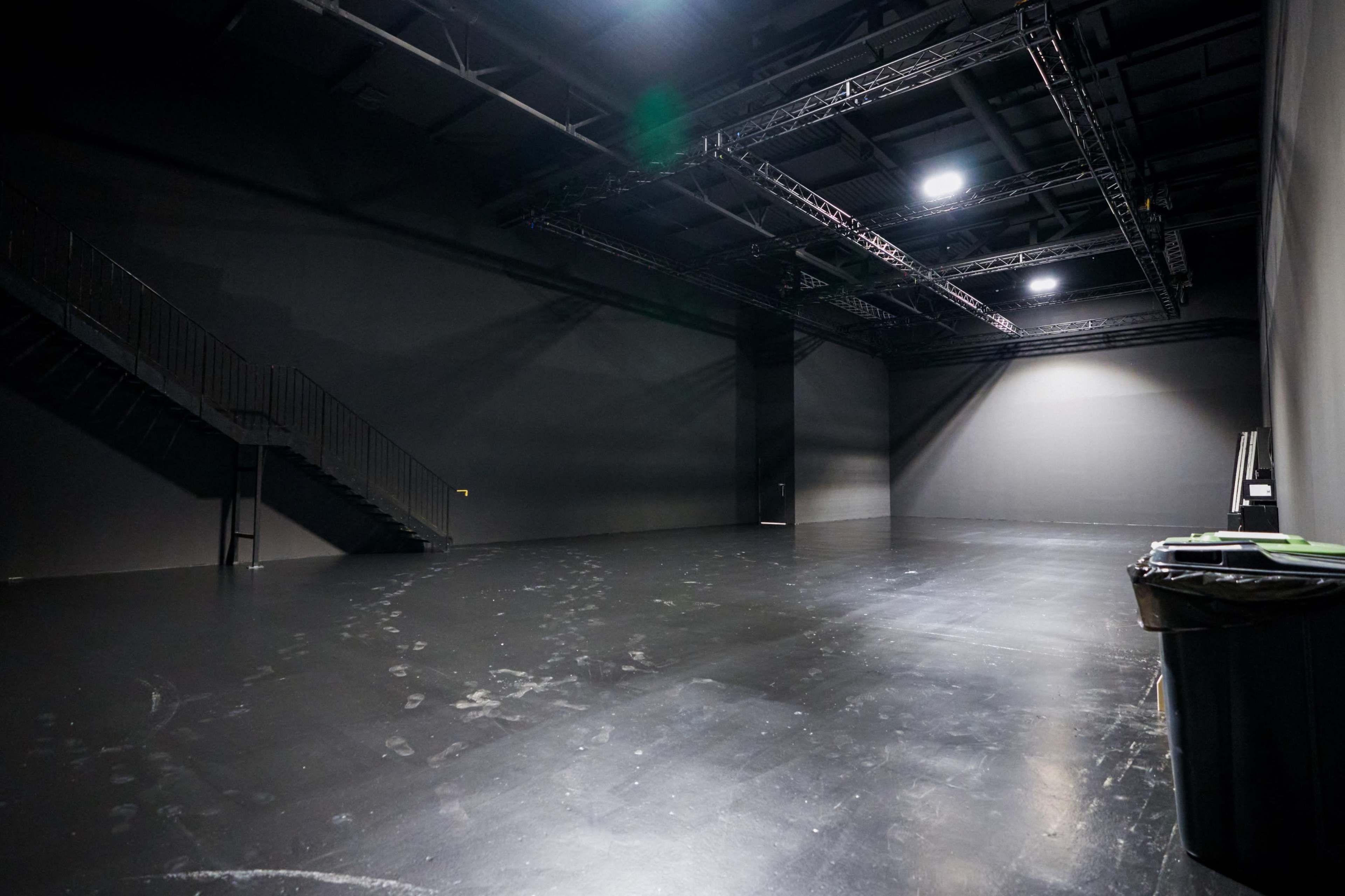 The image shows a large, empty studio space with a black floor, a staircase to the left, and a trash bin in the corner.