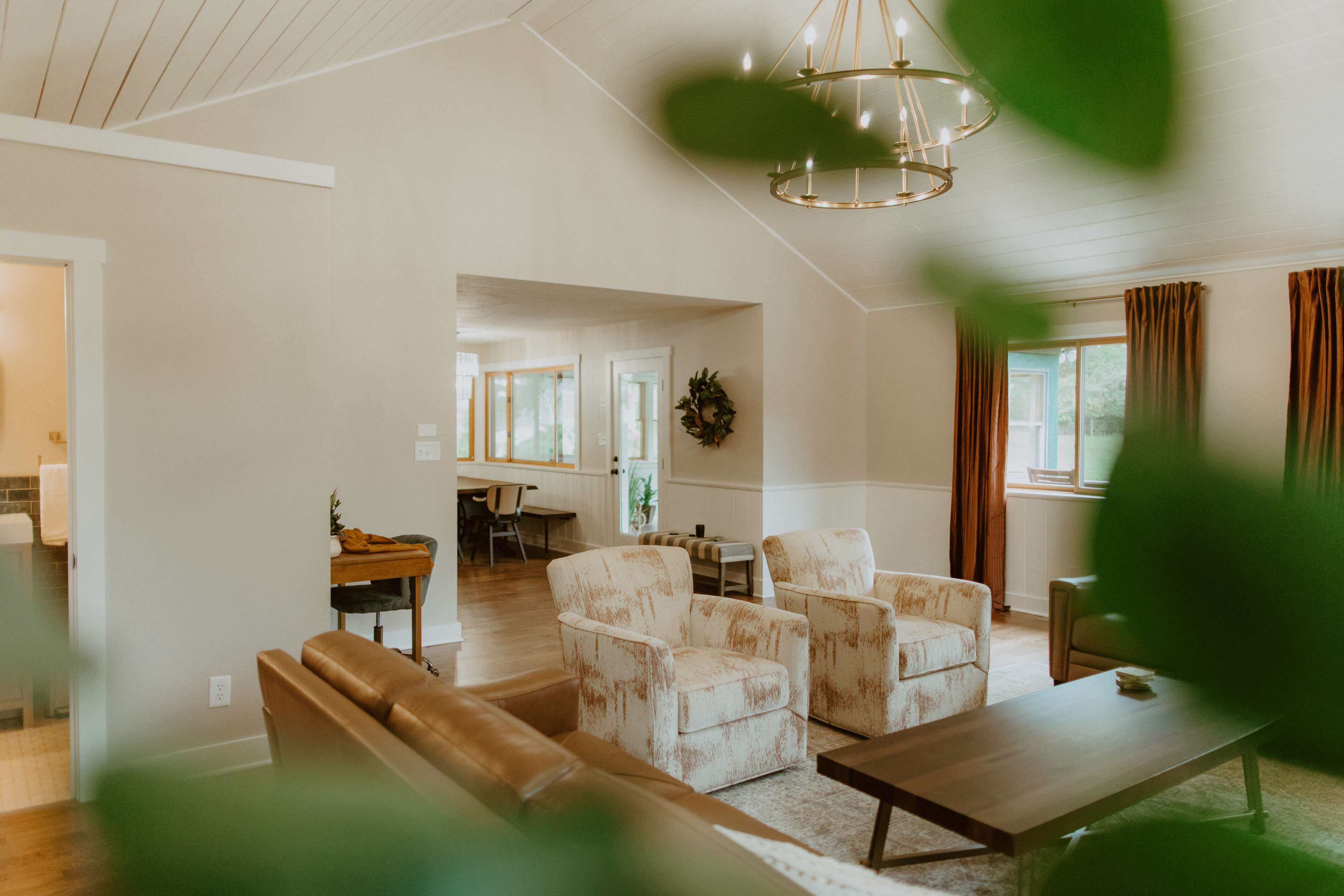 A cozy living room with two patterned chairs, a brown sofa, and a wooden coffee table, all beneath a chandelier, framed by large windows and plants.