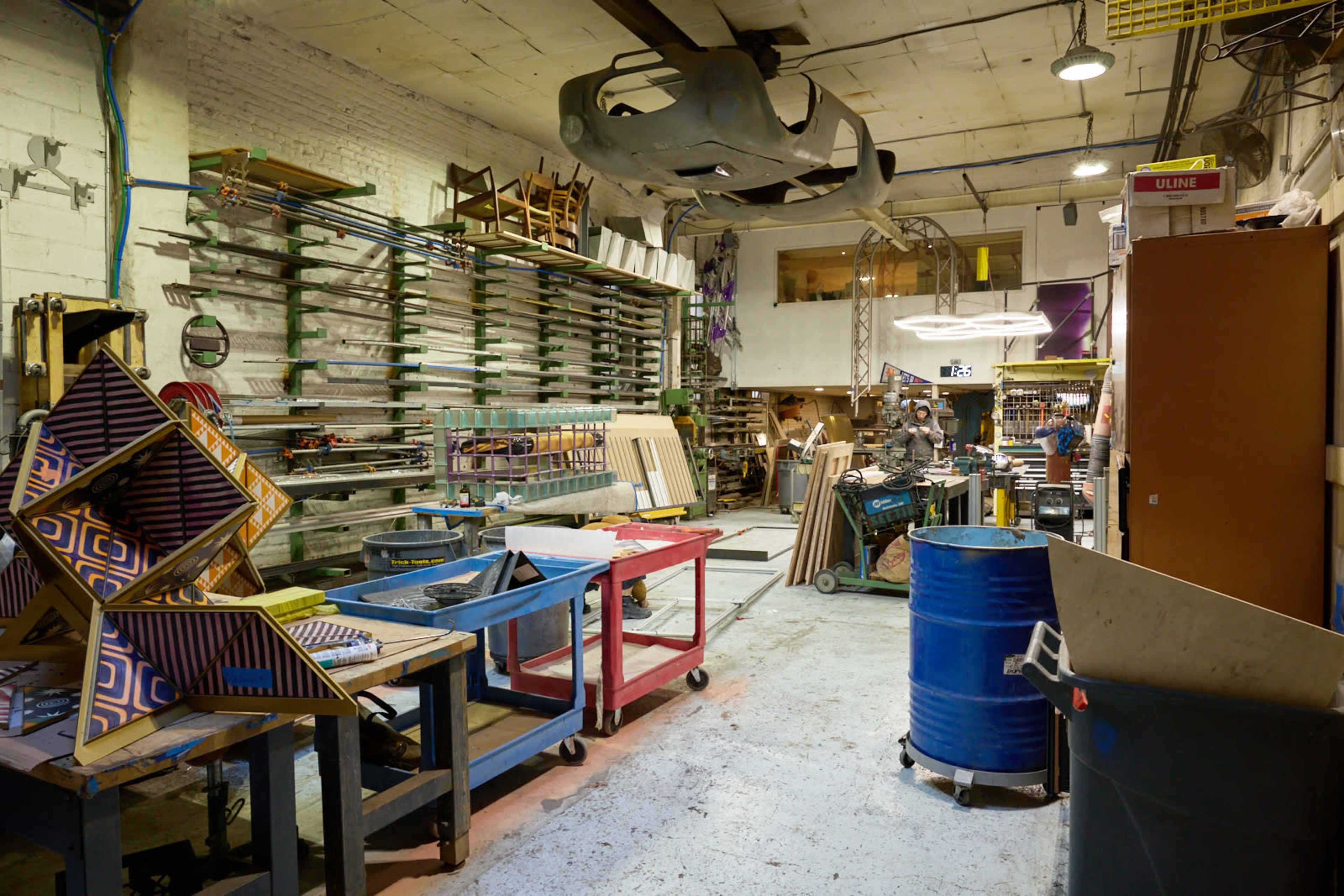 The image shows a spacious workshop filled with various tools, worktables, and materials organized along the walls, with a large overhead structure visible in the ceiling.