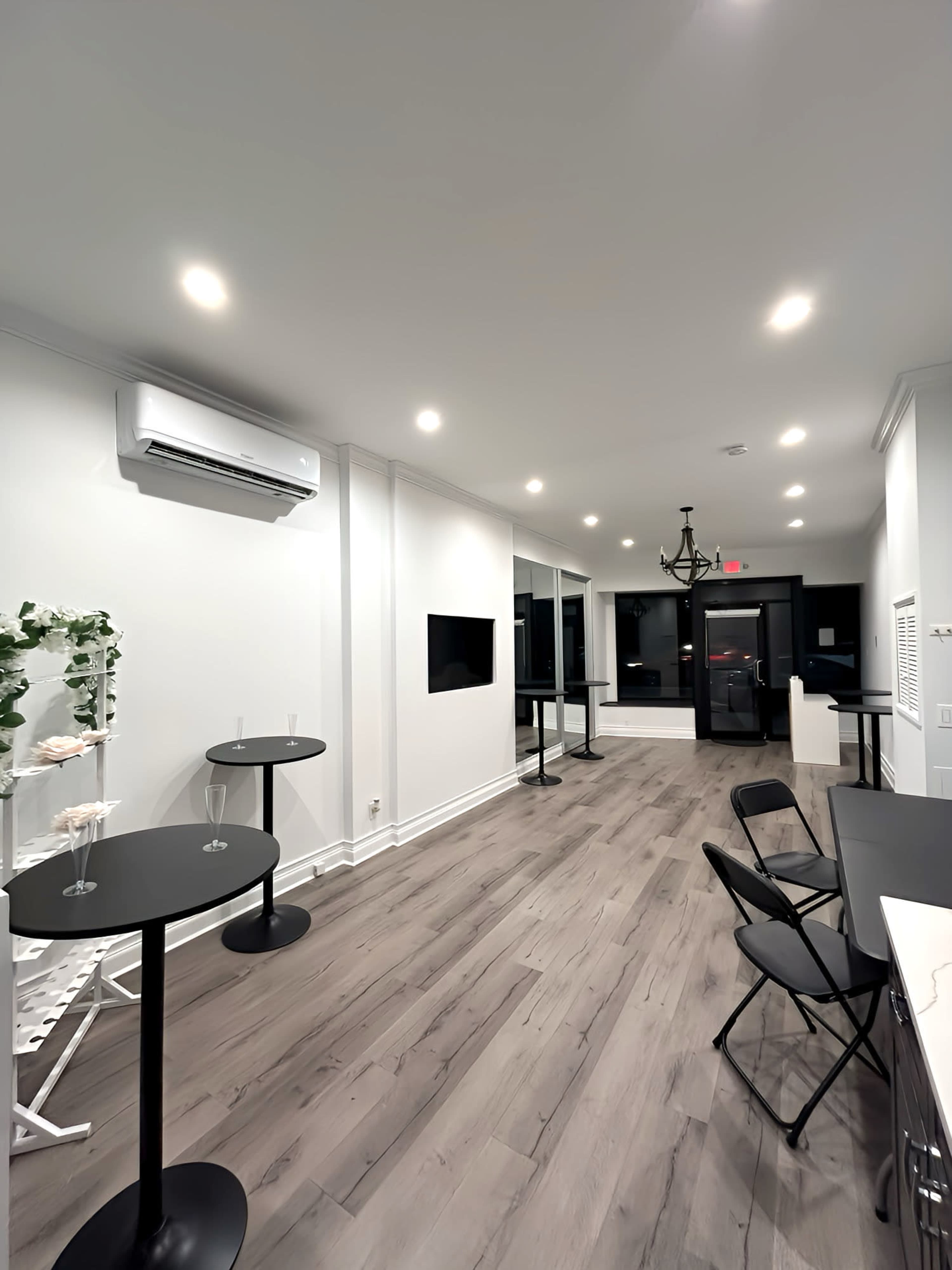 The image shows a modern indoor space with light-colored walls, wooden flooring, and several black tables and chairs arranged in the area.