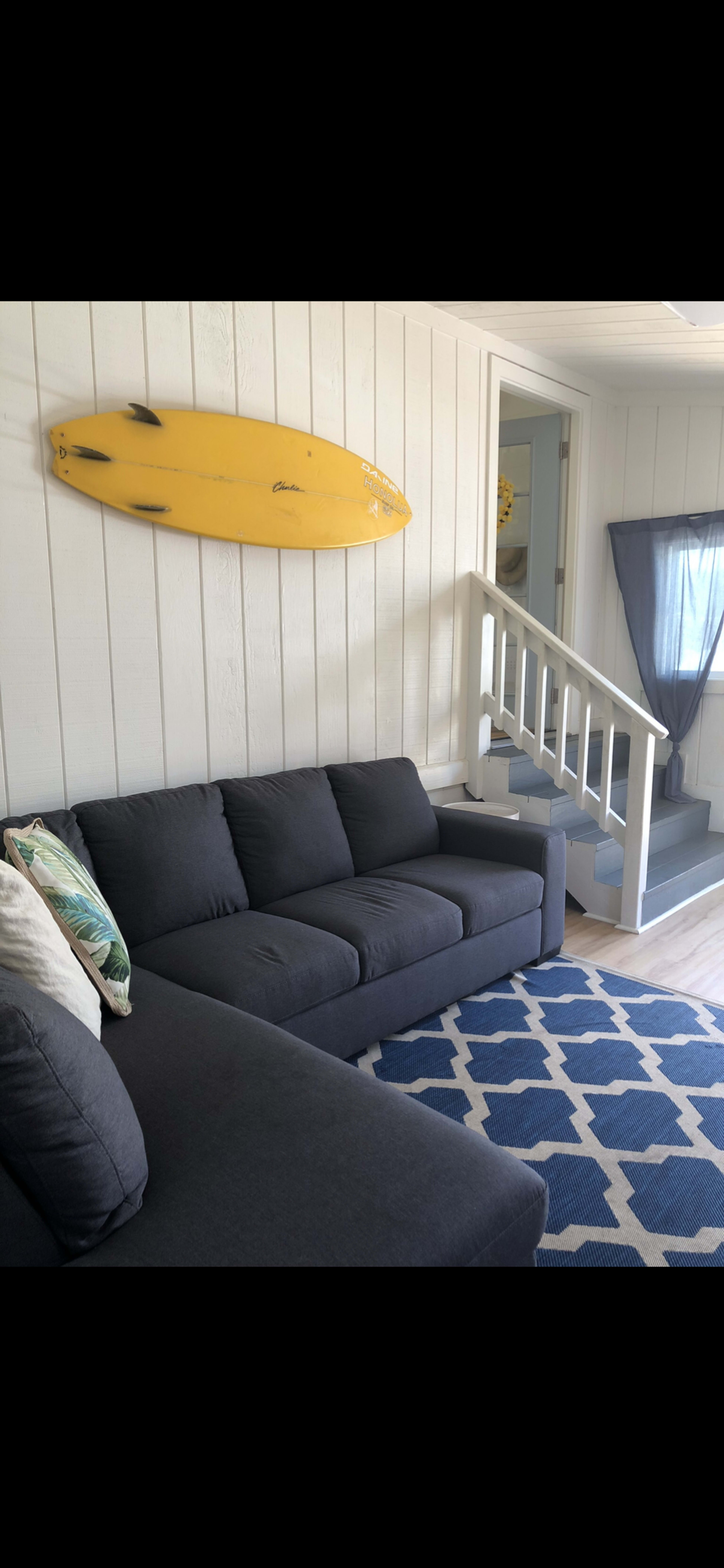 A modern living room with a dark gray sectional sofa, a blue and white patterned rug, and a yellow surfboard mounted on the white shiplap wall.