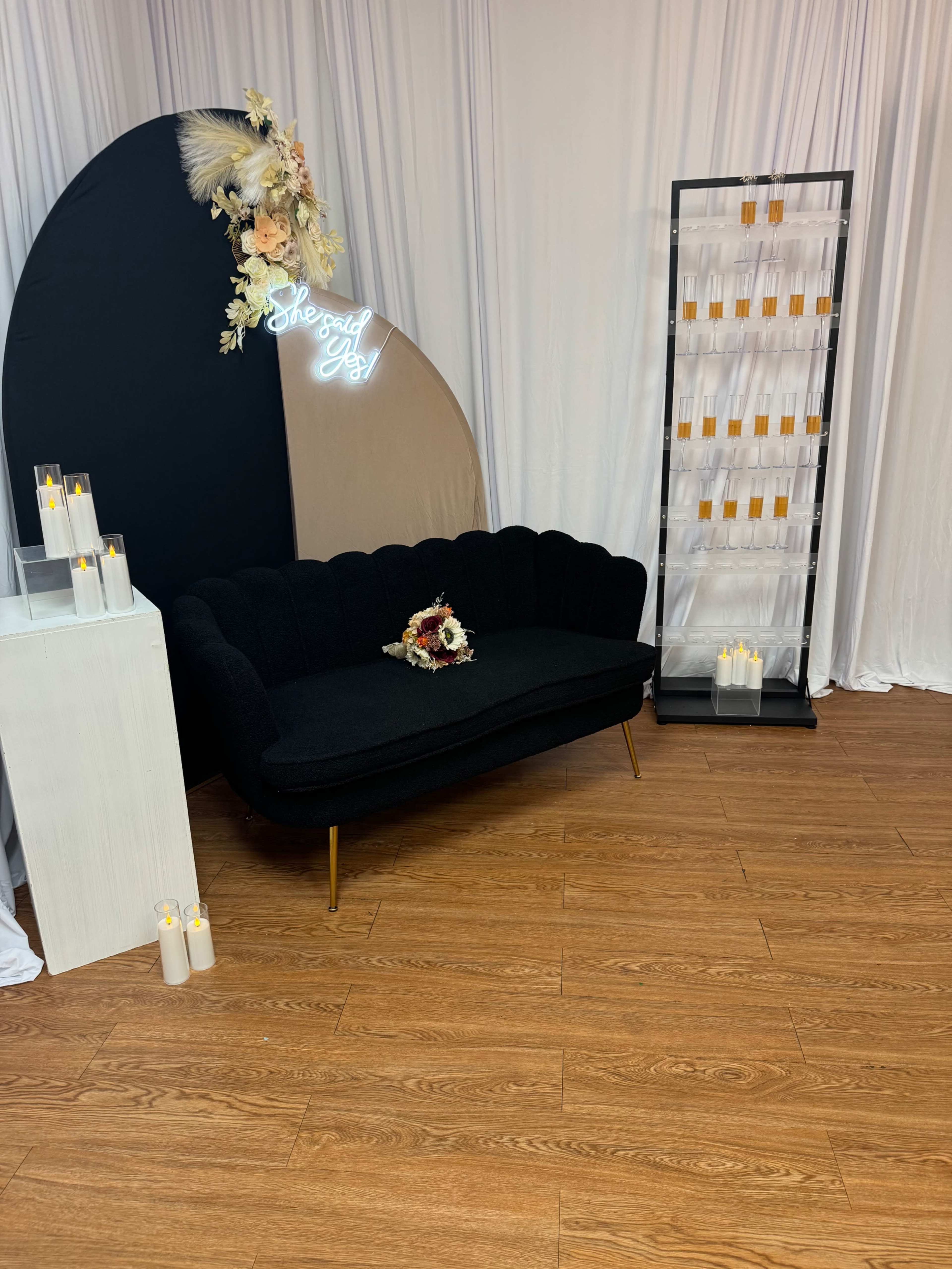 The image shows a black tufted couch placed against a backdrop curtain, accompanied by a floral arrangement and a display of candles on a stand.