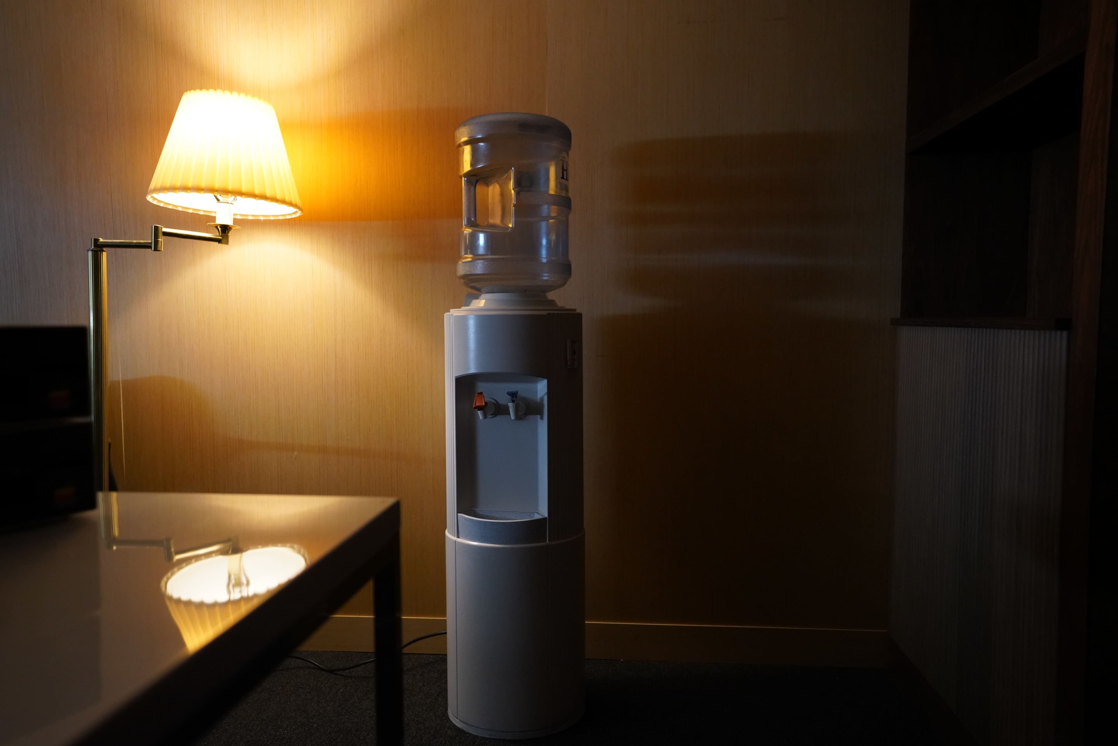 A water cooler stands beside a lamp in a dimly lit room with a wooden shelf in the background.