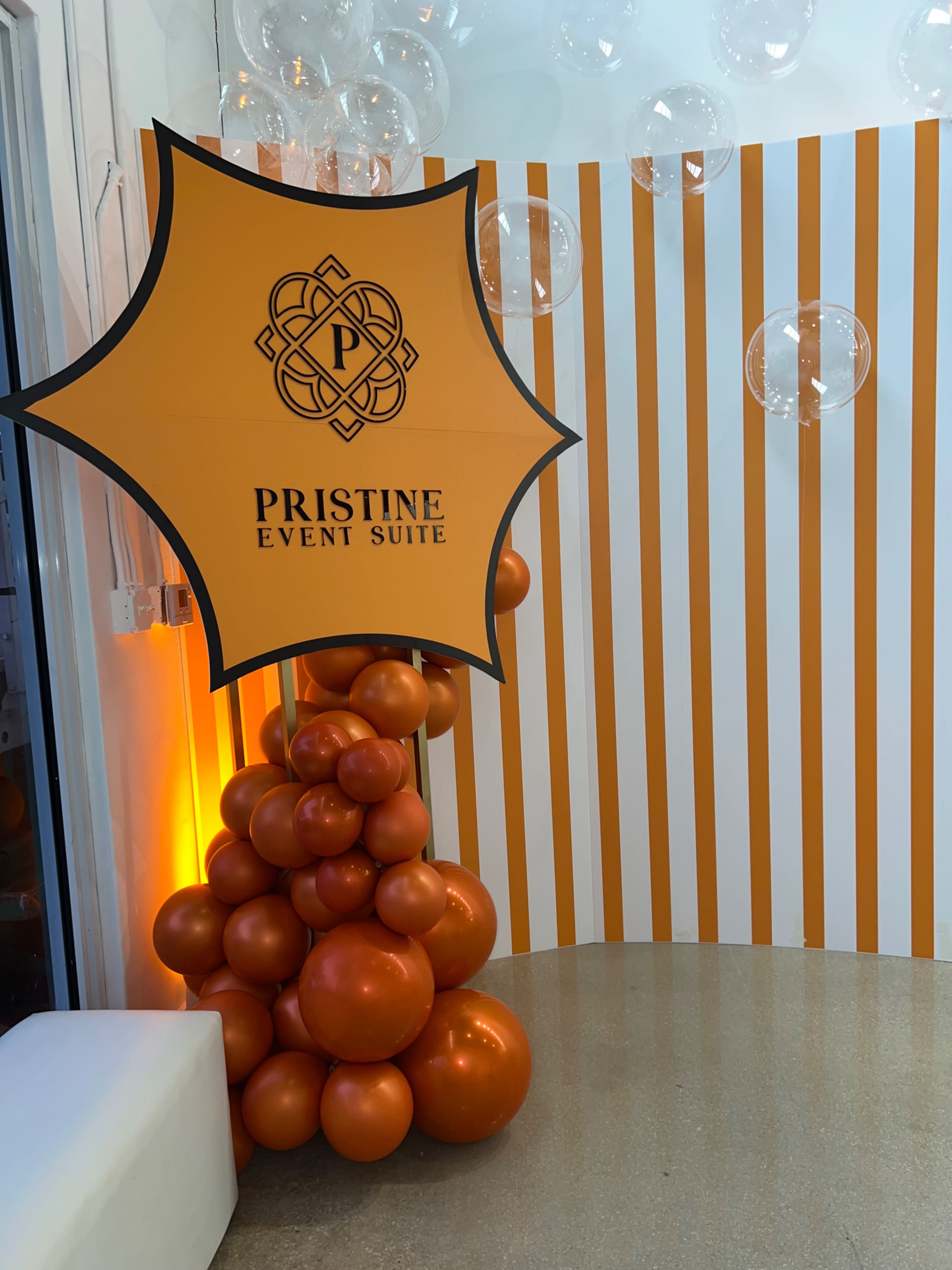 The image features a colorful event space decorated with an orange and white striped wall and a cluster of orange balloons supporting a sign that reads "Pristine Event Suite."