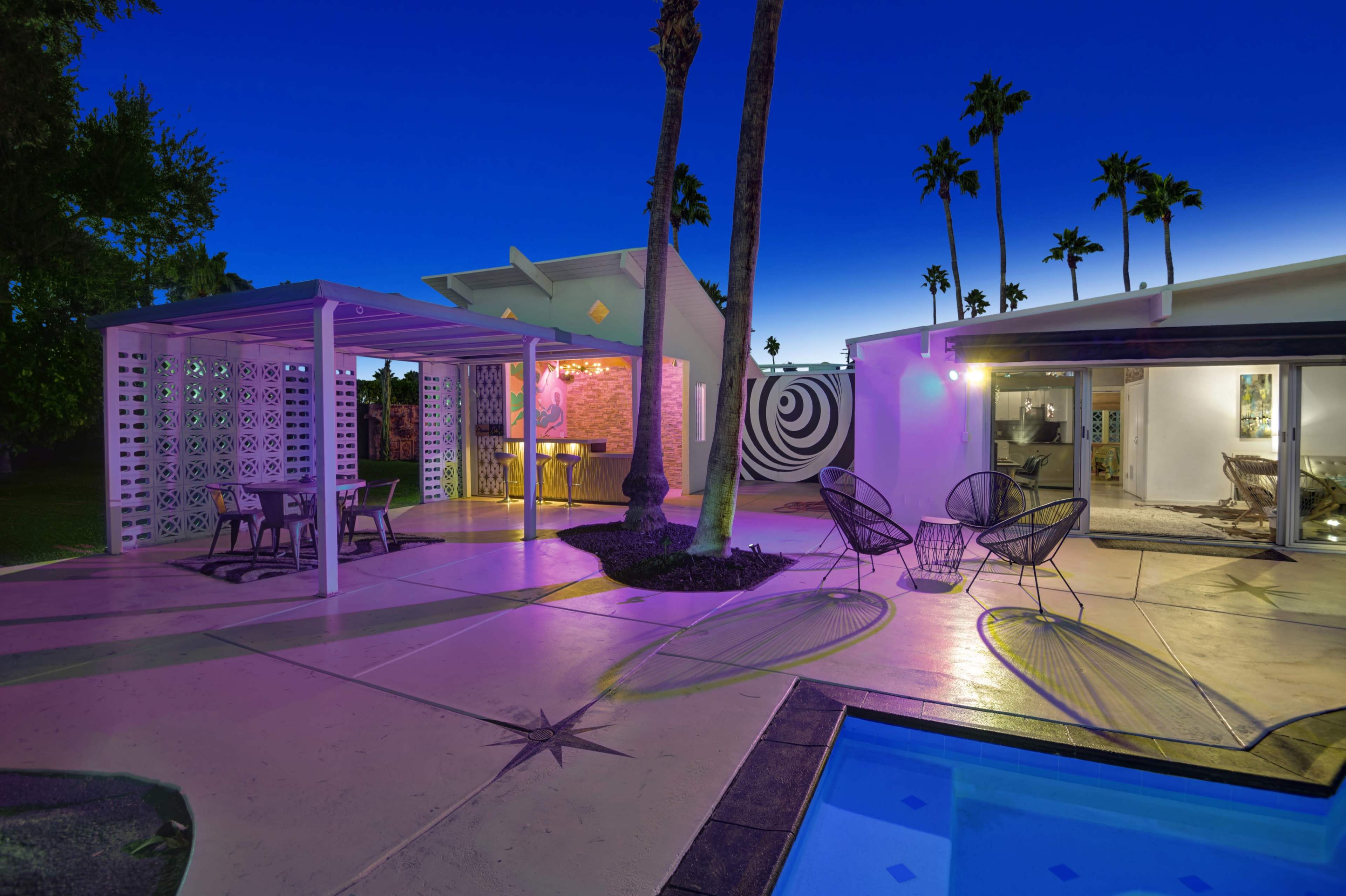 The image shows a modern backyard with a swimming pool, outdoor seating area, and a house featuring geometric patterns at dusk.