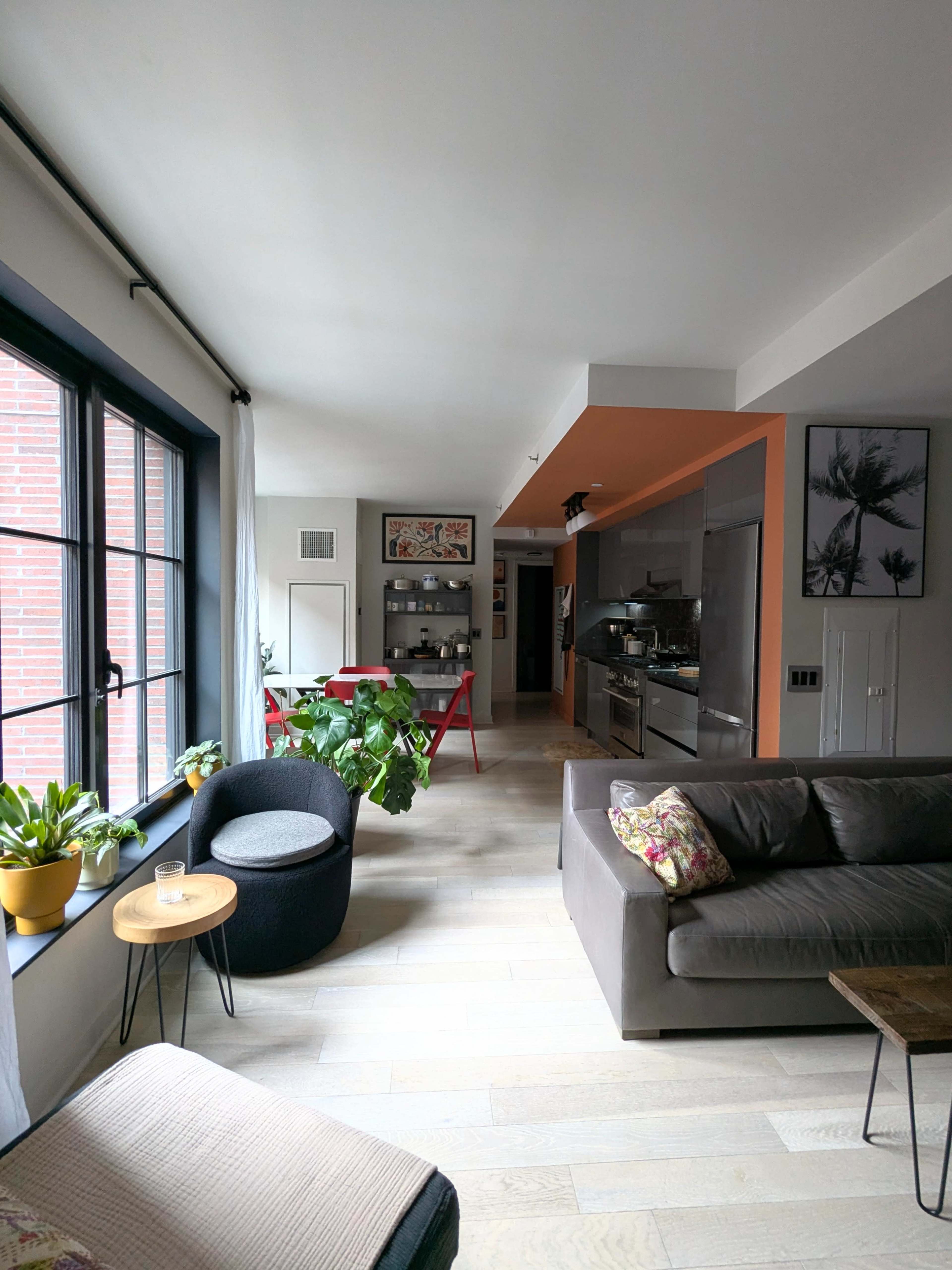 The image shows a modern living space with a cozy seating area, large windows, and an open kitchen in the background.