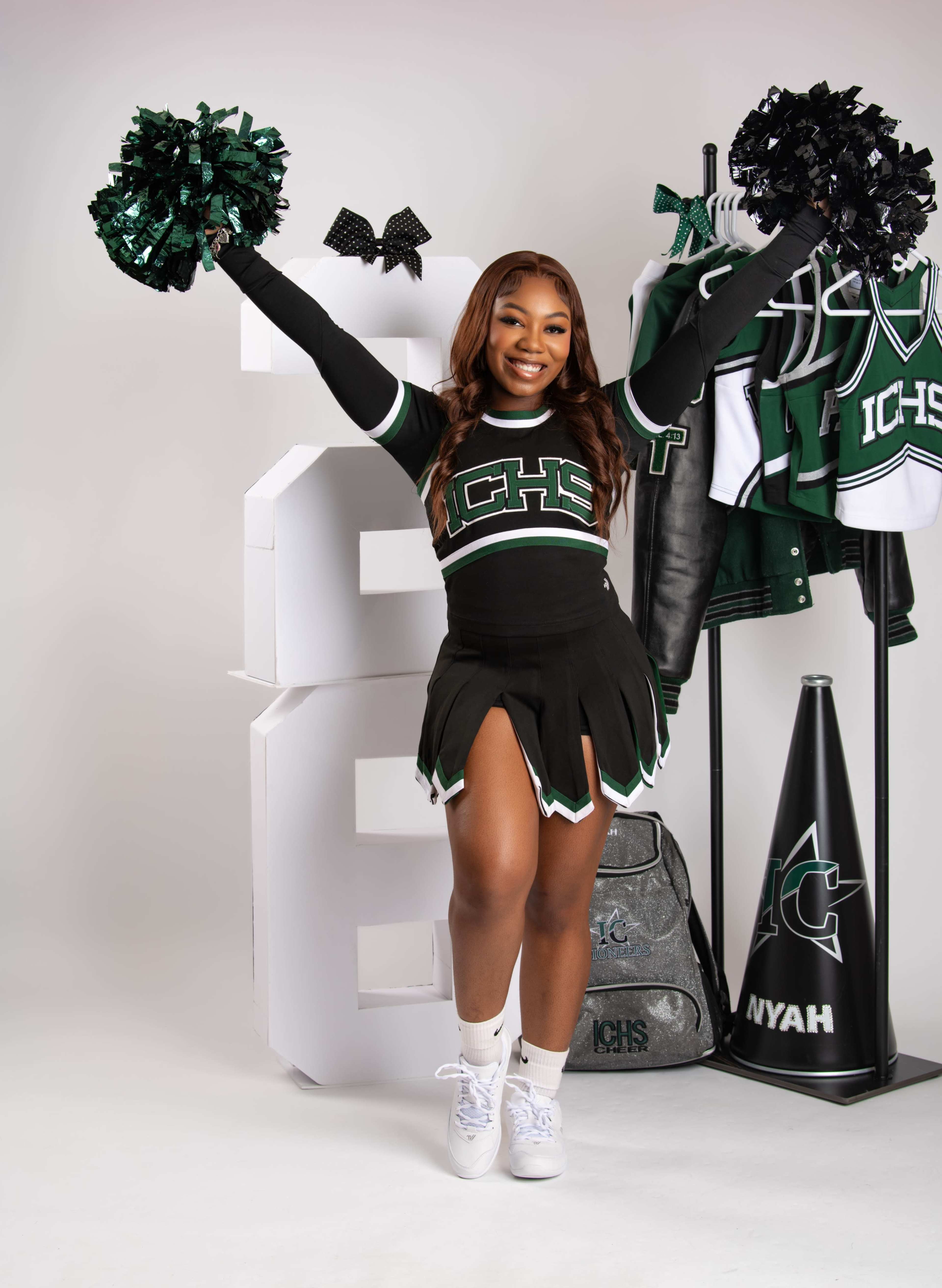 A cheerleader poses with pom-poms in front of a large letter, wearing her team's uniform and surrounded by cheerleading gear.