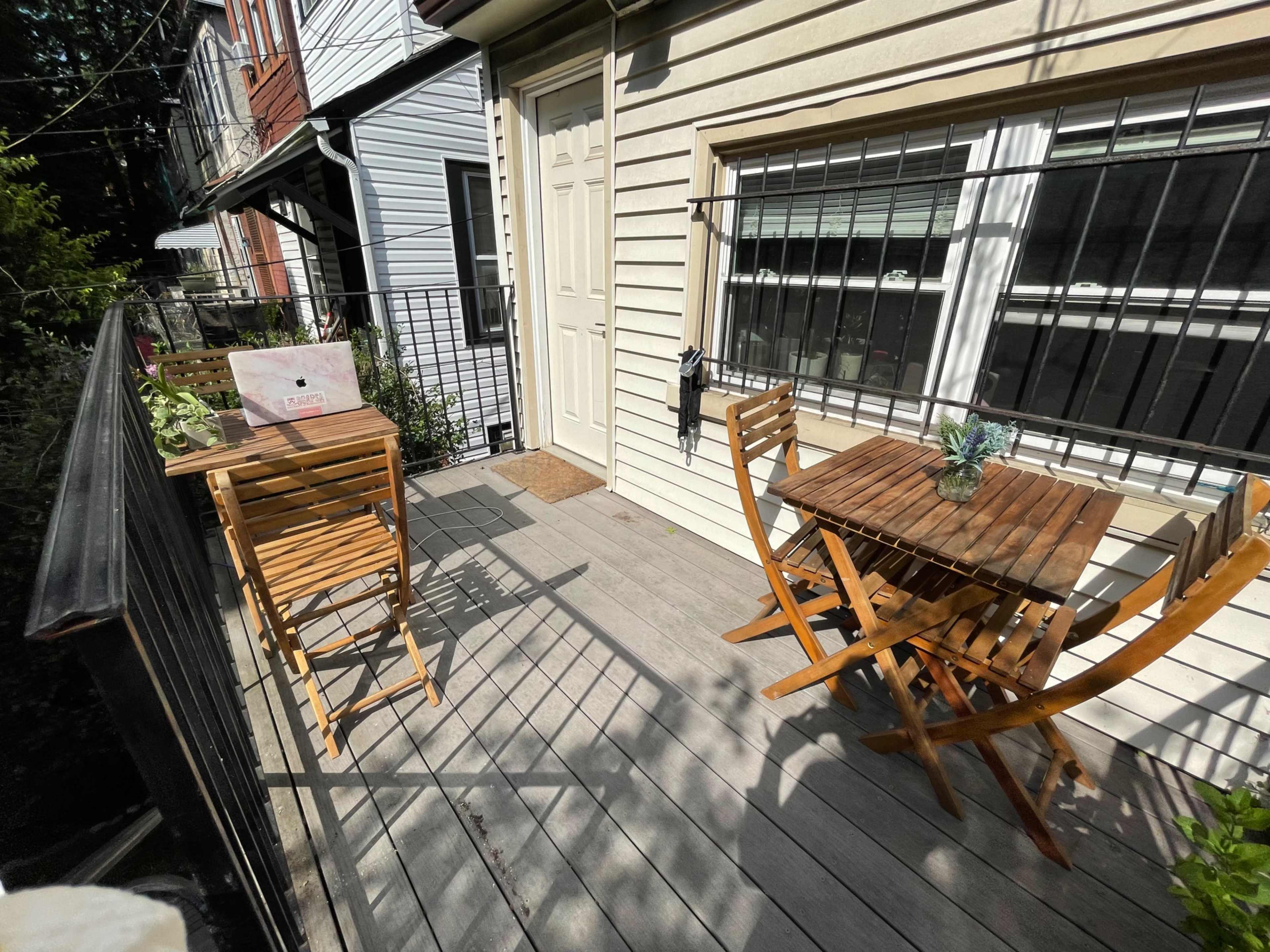 A small balcony with two wooden tables and chairs, surrounded by buildings and greenery.