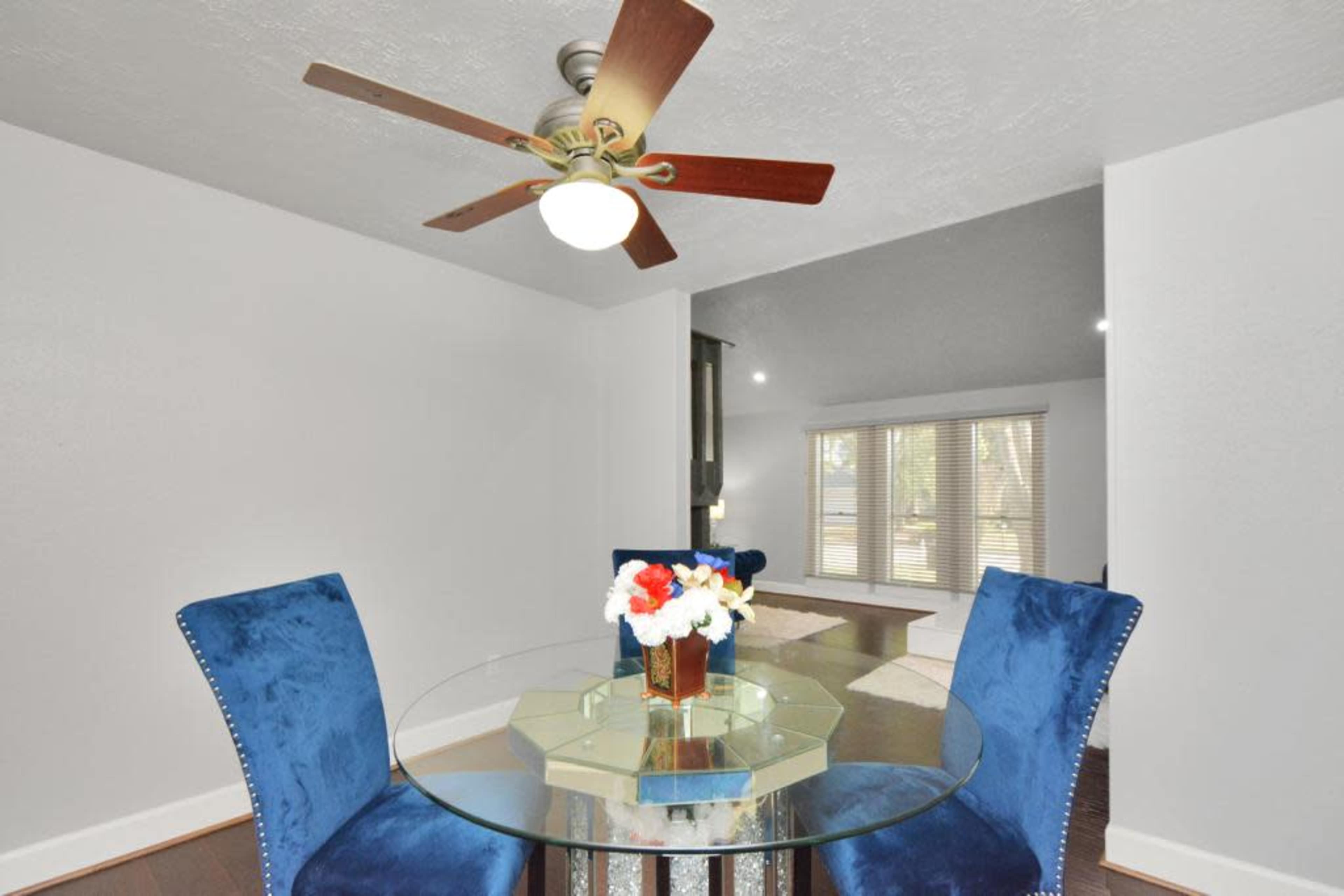 A glass dining table with blue chairs is positioned under a ceiling fan in a room with light-colored walls and a doorway leading to a living space with large windows.