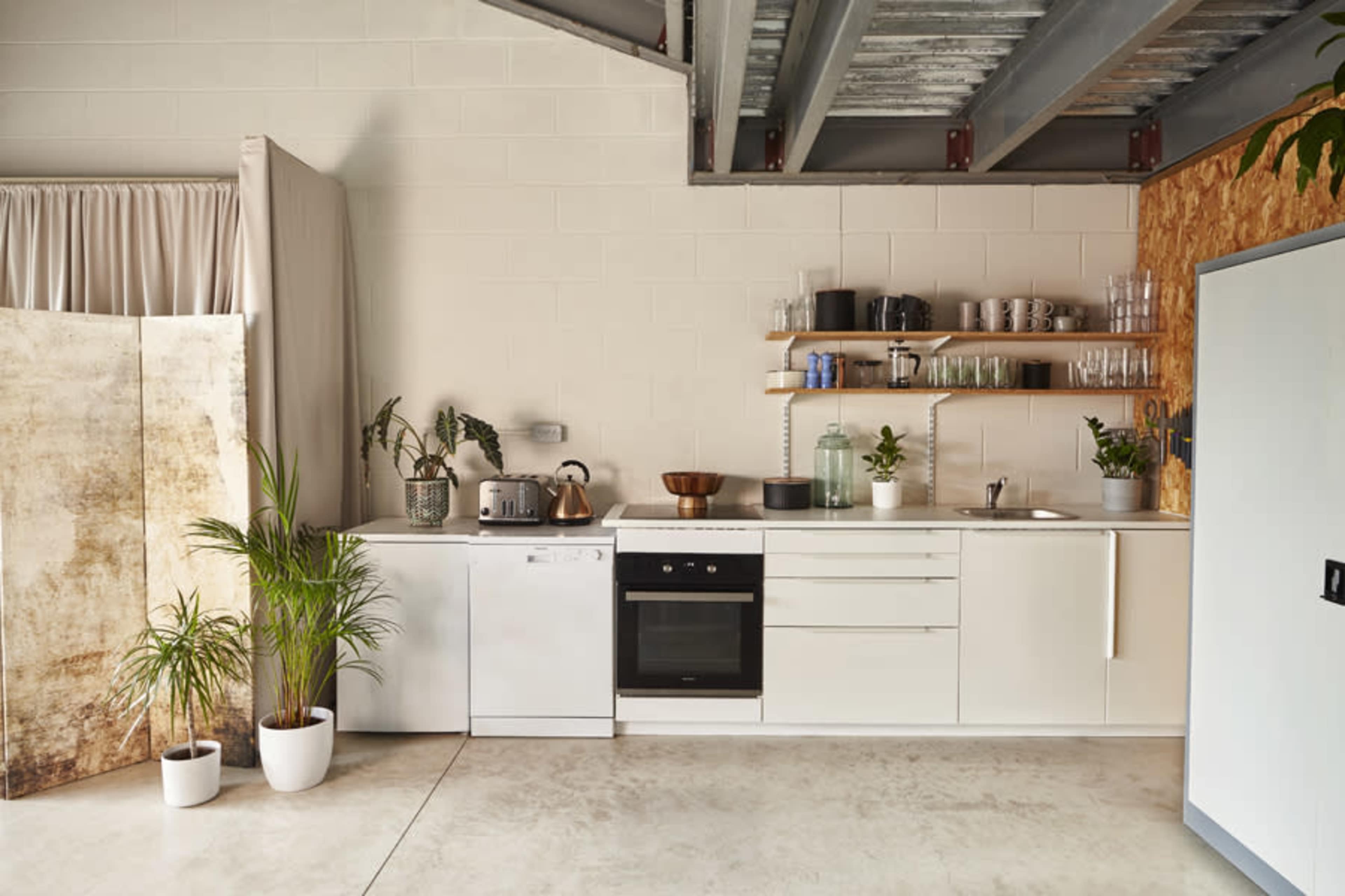 The image shows a modern kitchen with white cabinetry, a sink, a dishwasher, and shelves displaying glassware, set against a minimalist backdrop.