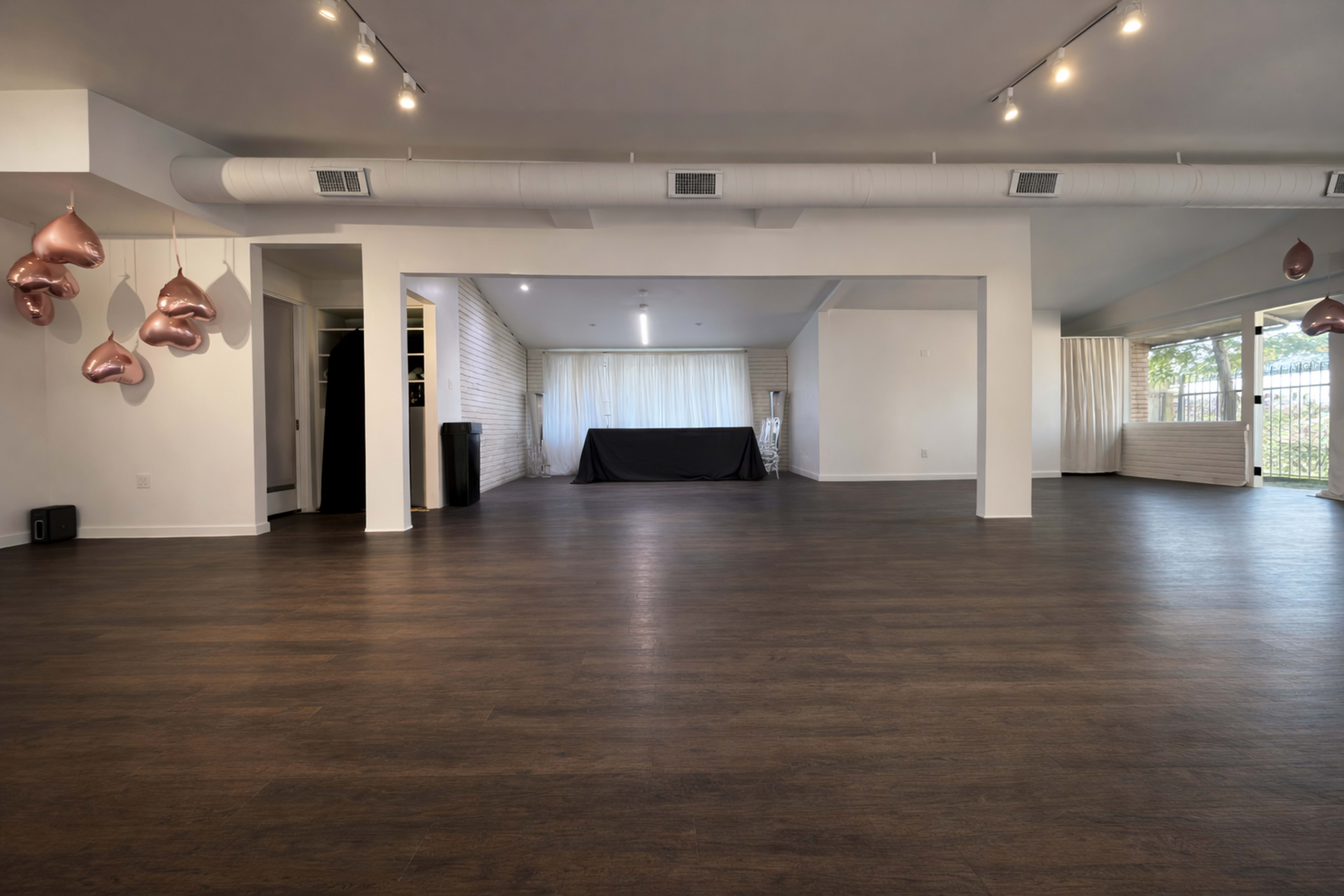 The image shows a spacious, empty room with wooden flooring, featuring a black table in the center, and several decorative heart-shaped balloons hanging from the ceiling.