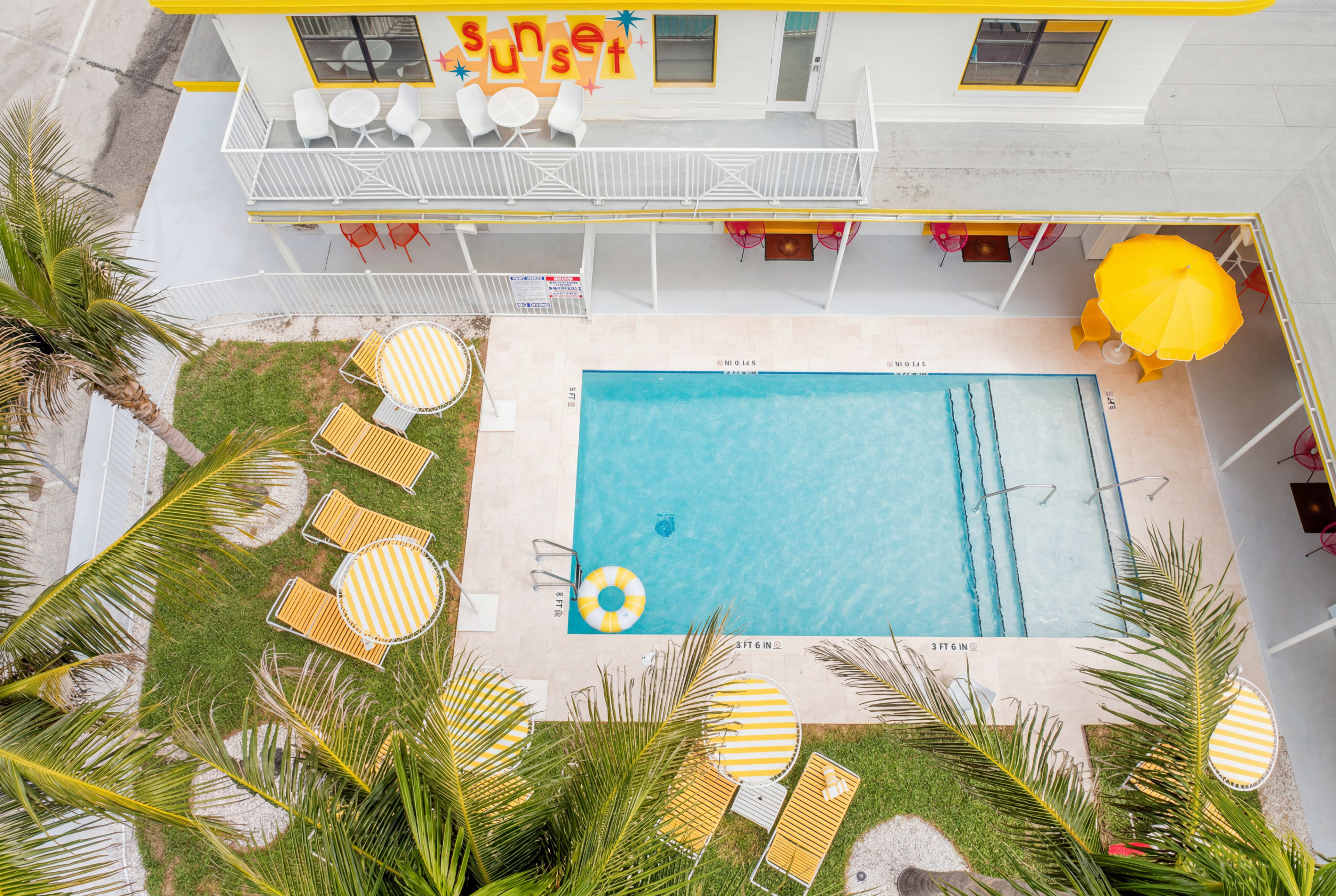 The image shows an outdoor pool area surrounded by palm trees, with striped lounge chairs and a yellow umbrella beside the clear water.