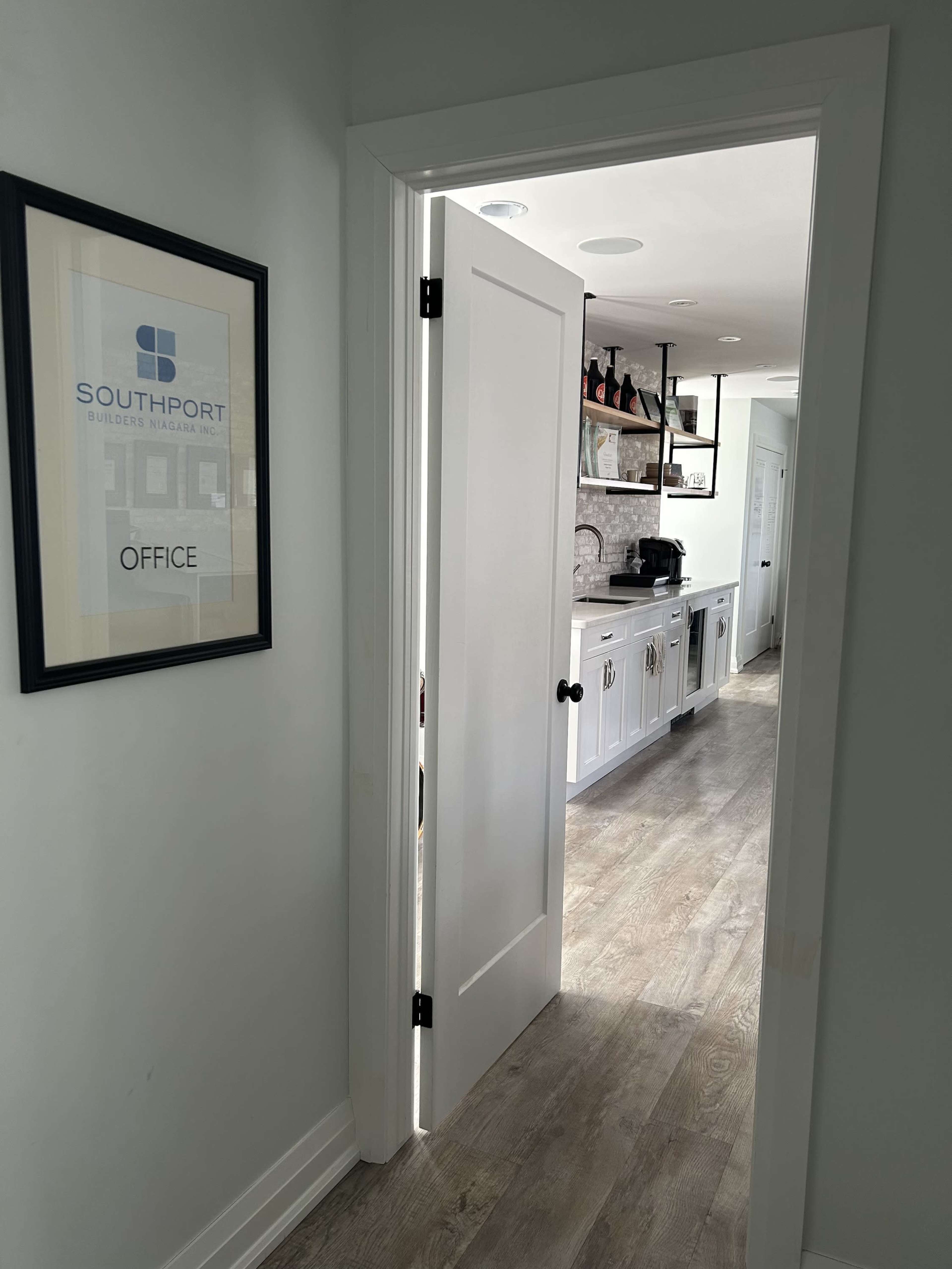 A partially open door leads to a well-lit office space with a view of a kitchen featuring cabinets and an espresso machine.